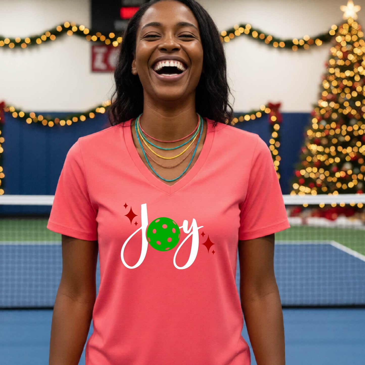 A woman smiling in a gym with a pickleball court  and a Christmas tree in the background, wearing a bright coral pink v-neck t-shirt with a 'Joy' graphic featuring a green pickleball and white script.
