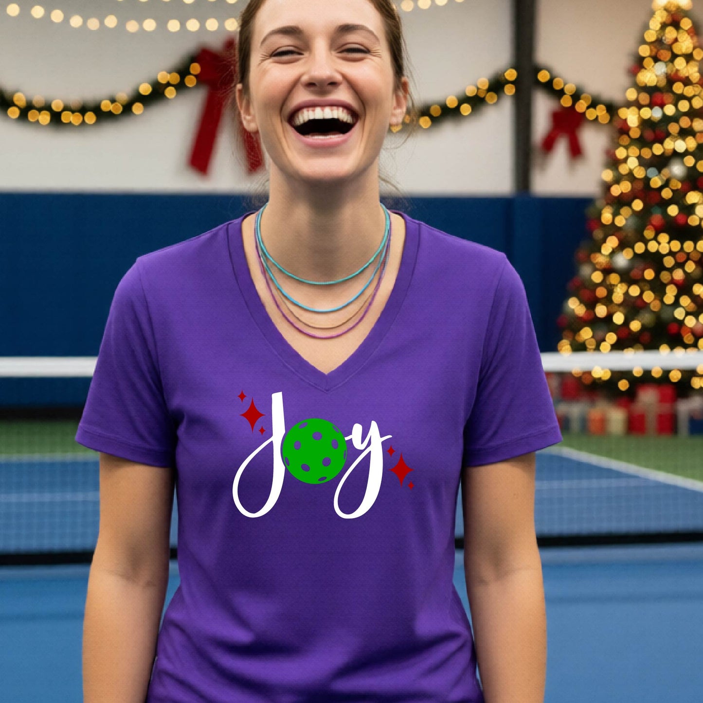 A woman smiling in a gym with a pickleball court  and a Christmas tree in the background, wearing a purple v-neck t-shirt with a 'Joy' graphic featuring a green pickleball and white script.