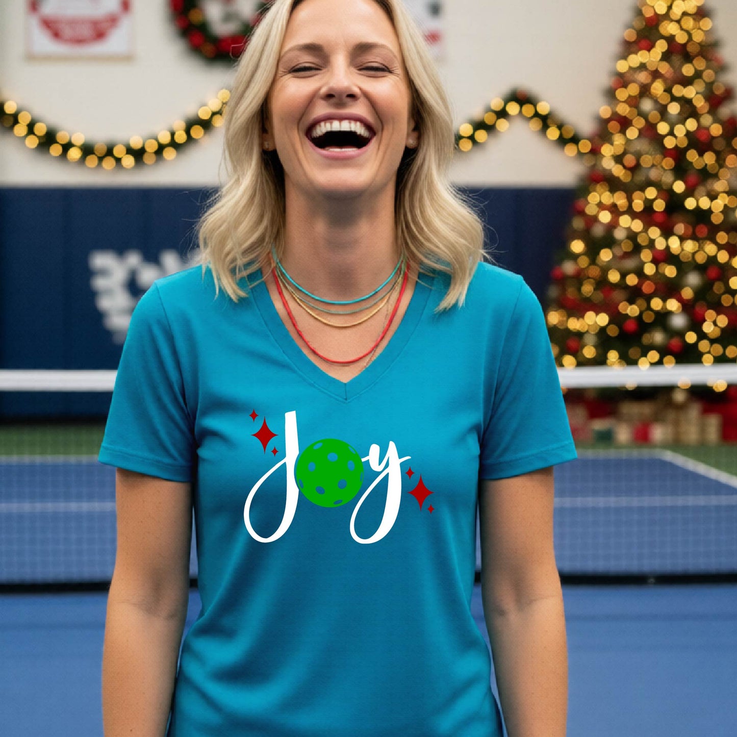 A woman smiling in a gym with a pickleball court  and a Christmas tree in the background, wearing a teal v-neck t-shirt with a 'Joy' graphic featuring a green pickleball and white script.