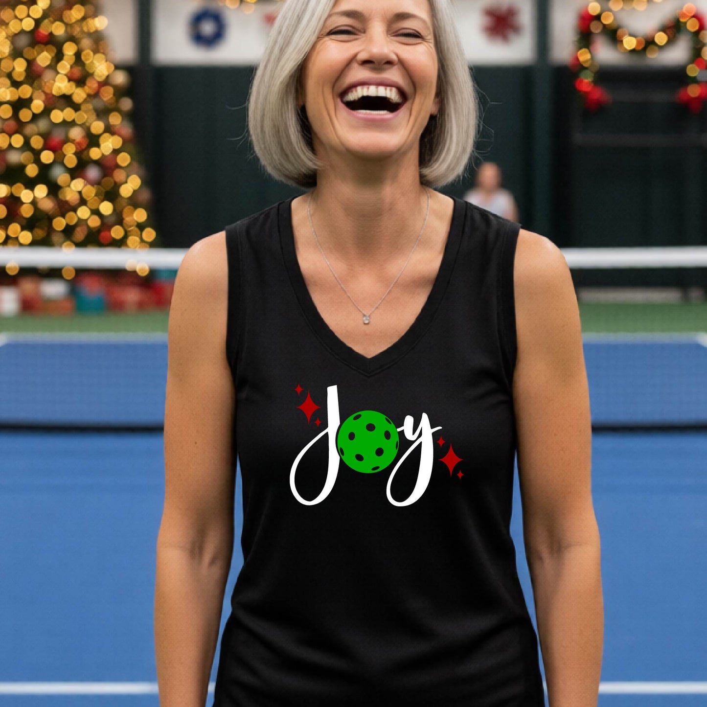 A woman laughing on a blue pickleball court wearing a black sleeveless tank top with a festive graphic that reads 'Joy' in white script, featuring a green pickleball and small red star accents. The background shows blurred Christmas decorations and lights. 