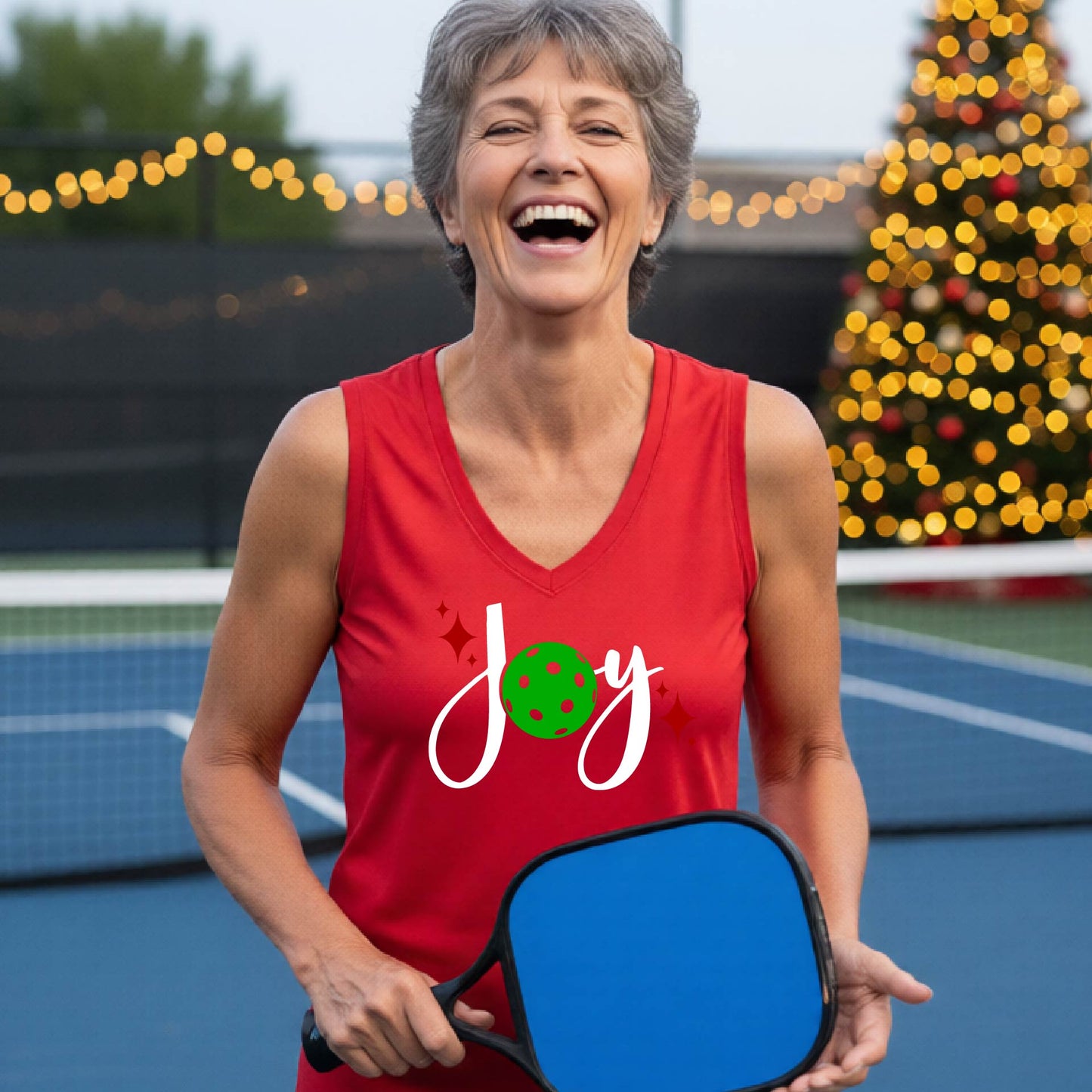 A woman laughing on a blue pickleball court wearing a red sleeveless tank top with a festive graphic that reads 'Joy' in white script, featuring a green pickleball and small red star accents. The background shows blurred Christmas decorations and lights. 