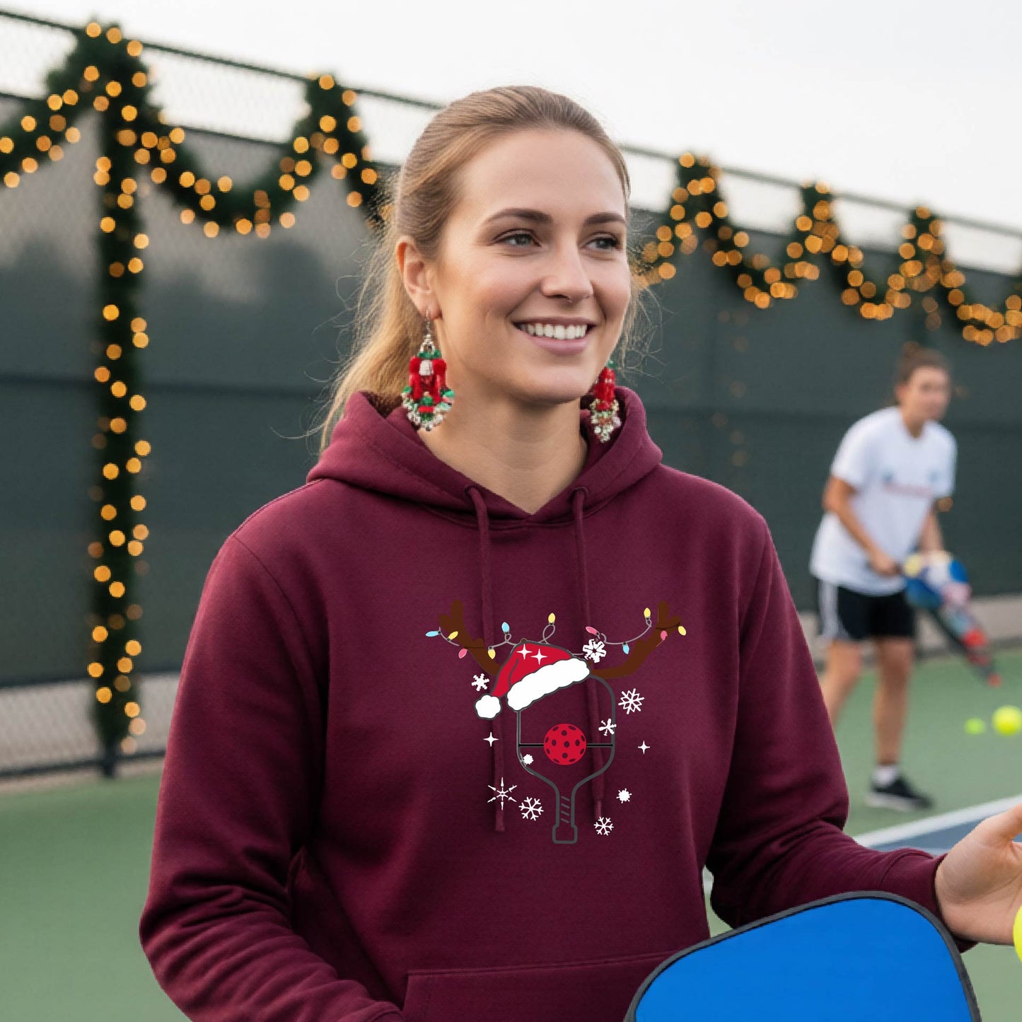 A smiling woman on an outdoor picleball court wears a maroon hoodie with a festive Christmas-themed pickleball padddle graphic on the front. The graphic features a pickleball paddle and ball decorated with a Santa hat and reindeer antlers.