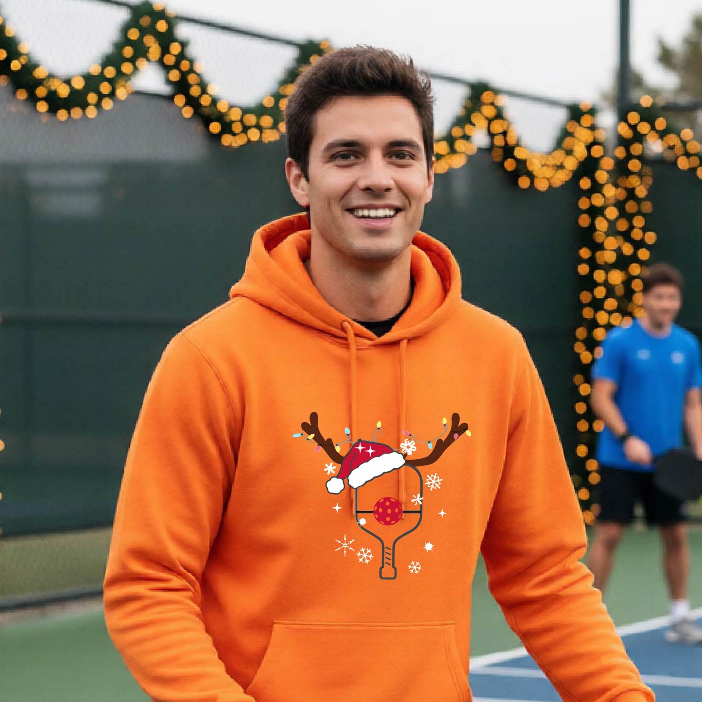 A smiling man on an outdoor picleball court wears a bright orange hoodie with a festive Christmas-themed pickleball padddle graphic on the front. The graphic features a pickleball paddle and ball decorated with a Santa hat and reindeer antlers.