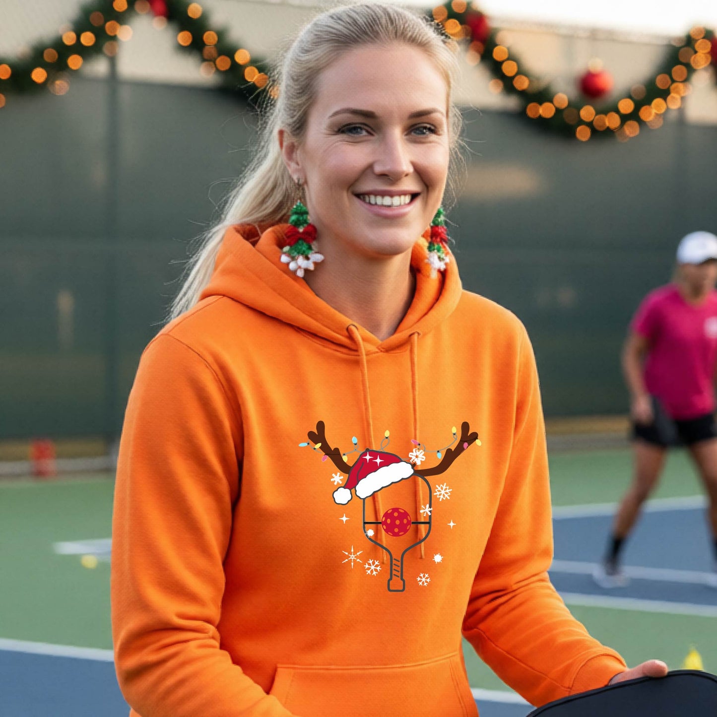A smiling woman on an outdoor picleball court wears a bright orange hoodie with a festive Christmas-themed pickleball padddle graphic on the front. The graphic features a pickleball paddle and ball decorated with a Santa hat and reindeer antlers.
