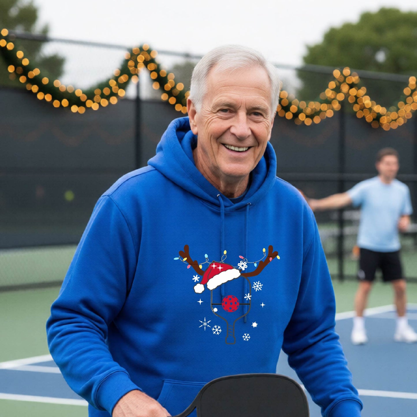 A smiling man on an outdoor picleball court wears a bright royal blue hoodie with a festive Christmas-themed pickleball padddle graphic on the front. The graphic features a pickleball paddle and ball decorated with a Santa hat and reindeer antlers.