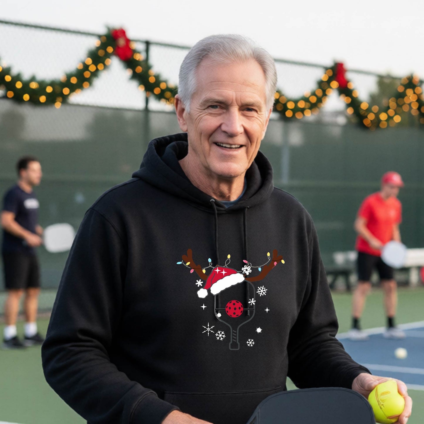 A smiling man on an outdoor picleball court wears a black hoodie with a festive Christmas-themed pickleball padddle graphic on the front. The graphic features a pickleball paddle and ball decorated with a Santa hat and reindeer antlers.