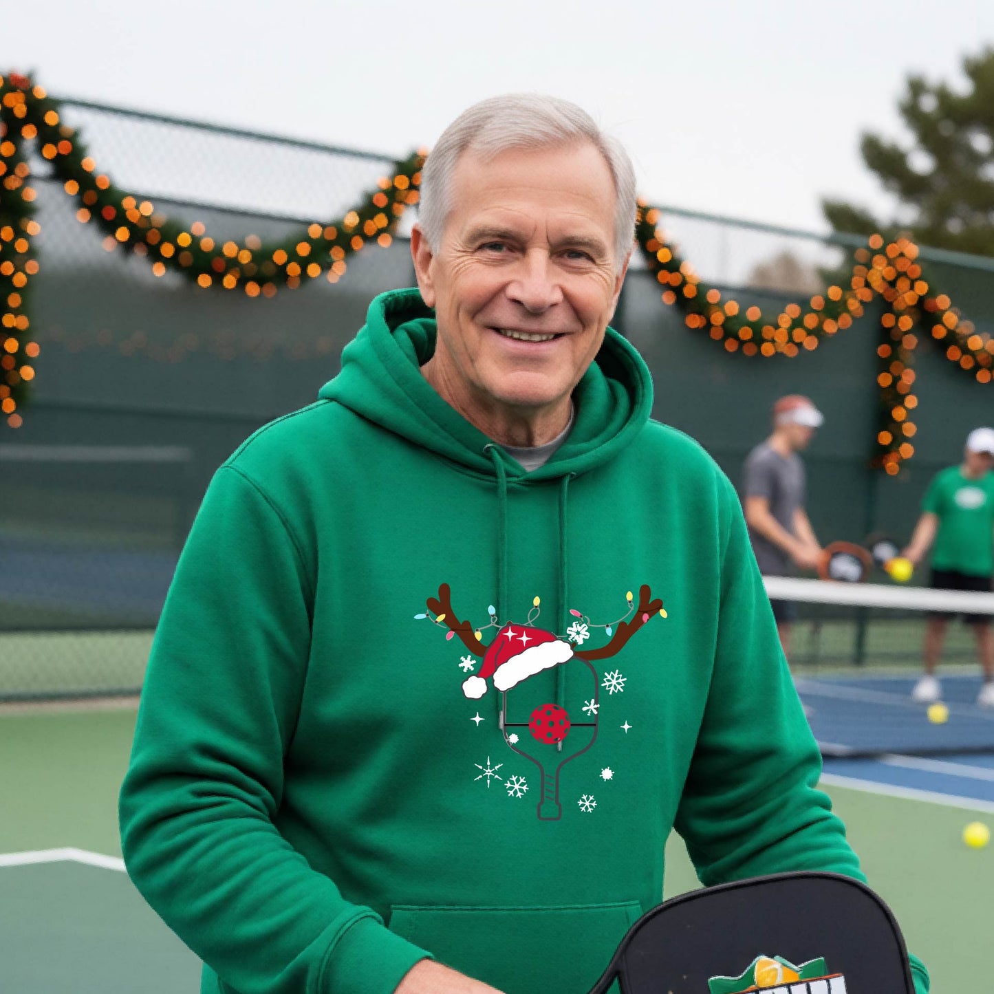 A smiling man on an outdoor picleball court wears a green hoodie with a festive Christmas-themed pickleball padddle graphic on the front. The graphic features a pickleball paddle and ball decorated with a Santa hat and reindeer antlers.