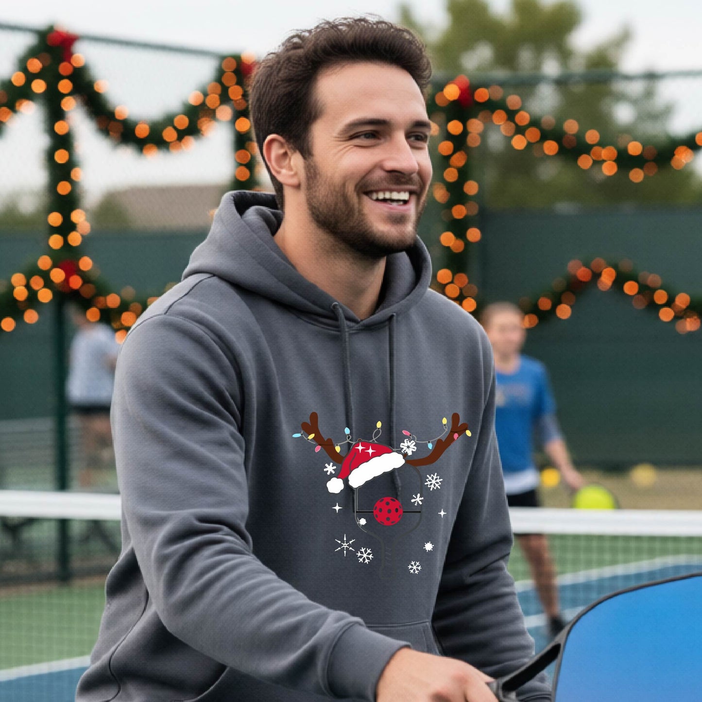A smiling man on an outdoor picleball court wears a gray hoodie with a festive Christmas-themed pickleball padddle graphic on the front. The graphic features a pickleball paddle and ball decorated with a Santa hat and reindeer antlers.