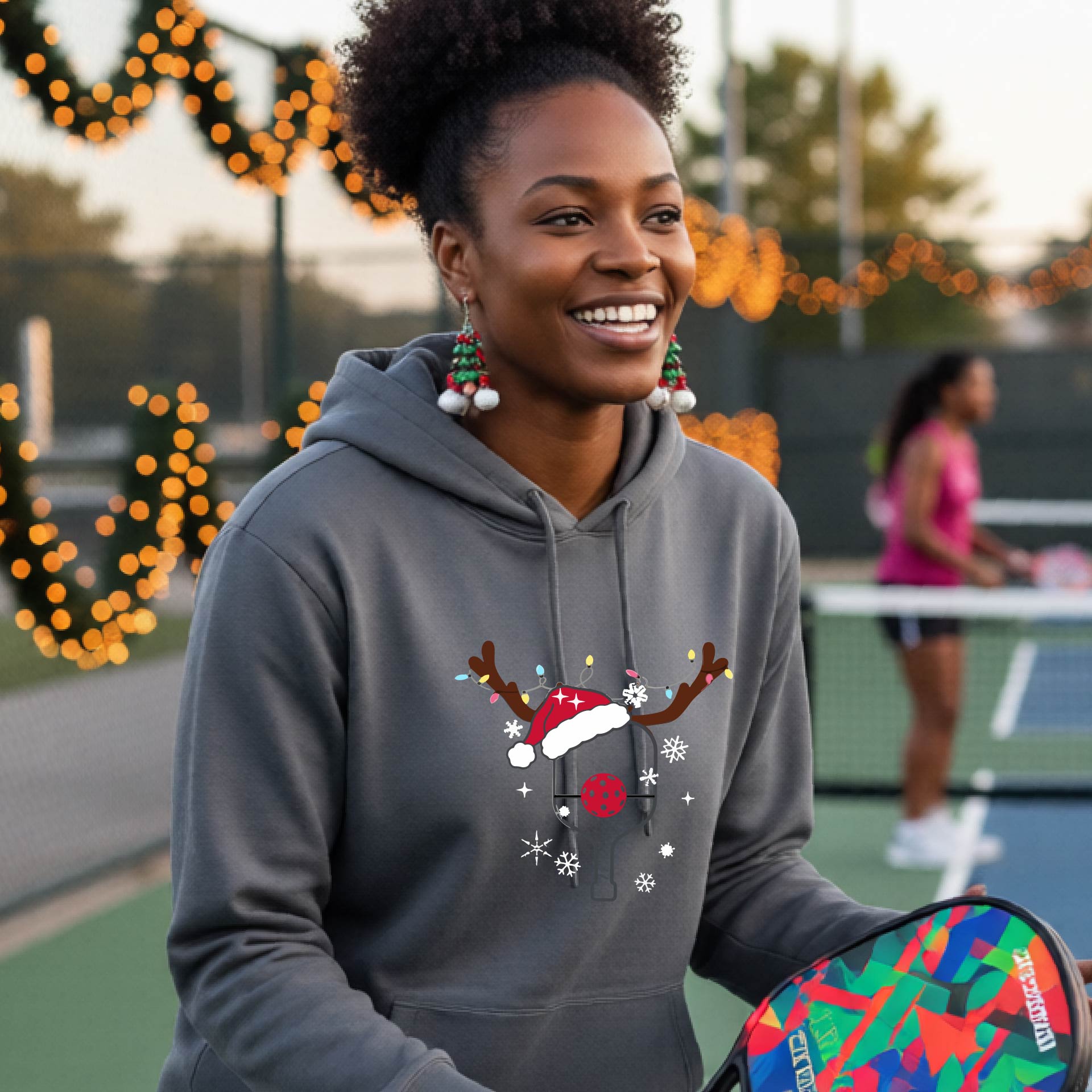 A smiling woman on an outdoor picleball court wears a gray hoodie with a festive Christmas-themed pickleball padddle graphic on the front. The graphic features a pickleball paddle and ball decorated with a Santa hat and reindeer antlers.