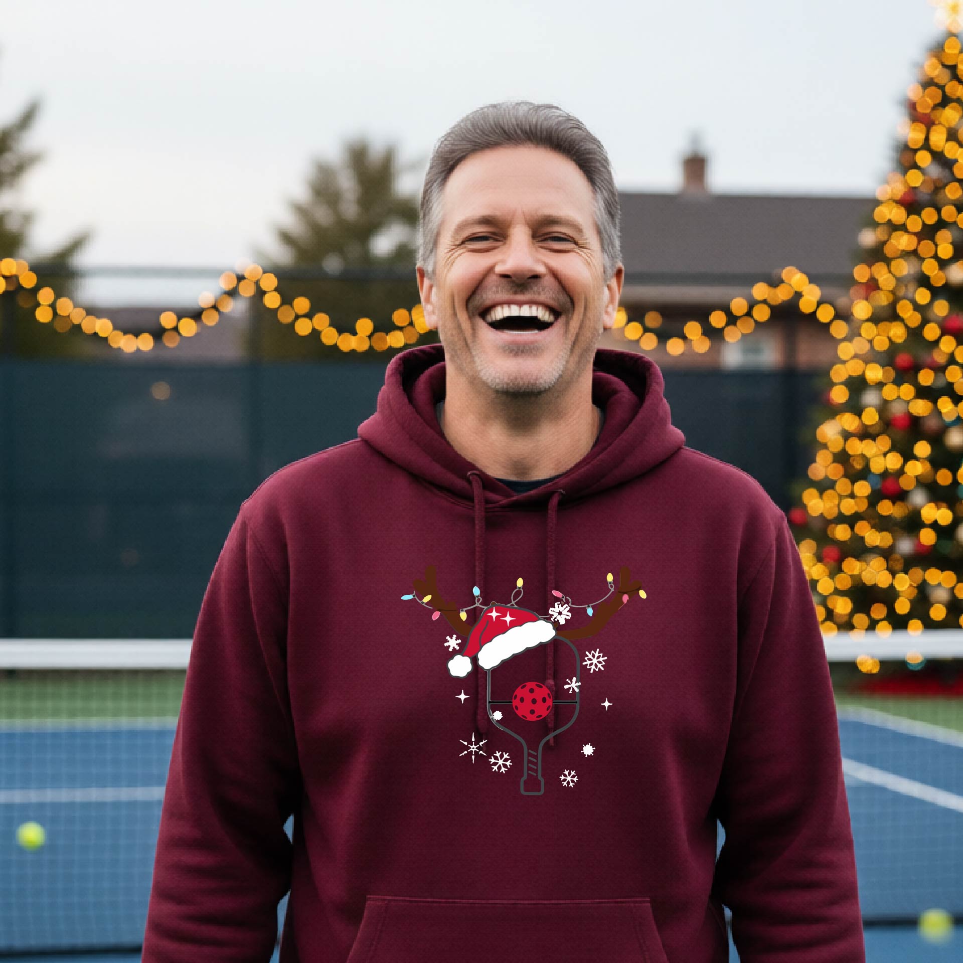 A smiling man on an outdoor picleball court wears a maroon hoodie with a festive Christmas-themed pickleball padddle graphic on the front. The graphic features a pickleball paddle and ball decorated with a Santa hat and reindeer antlers.