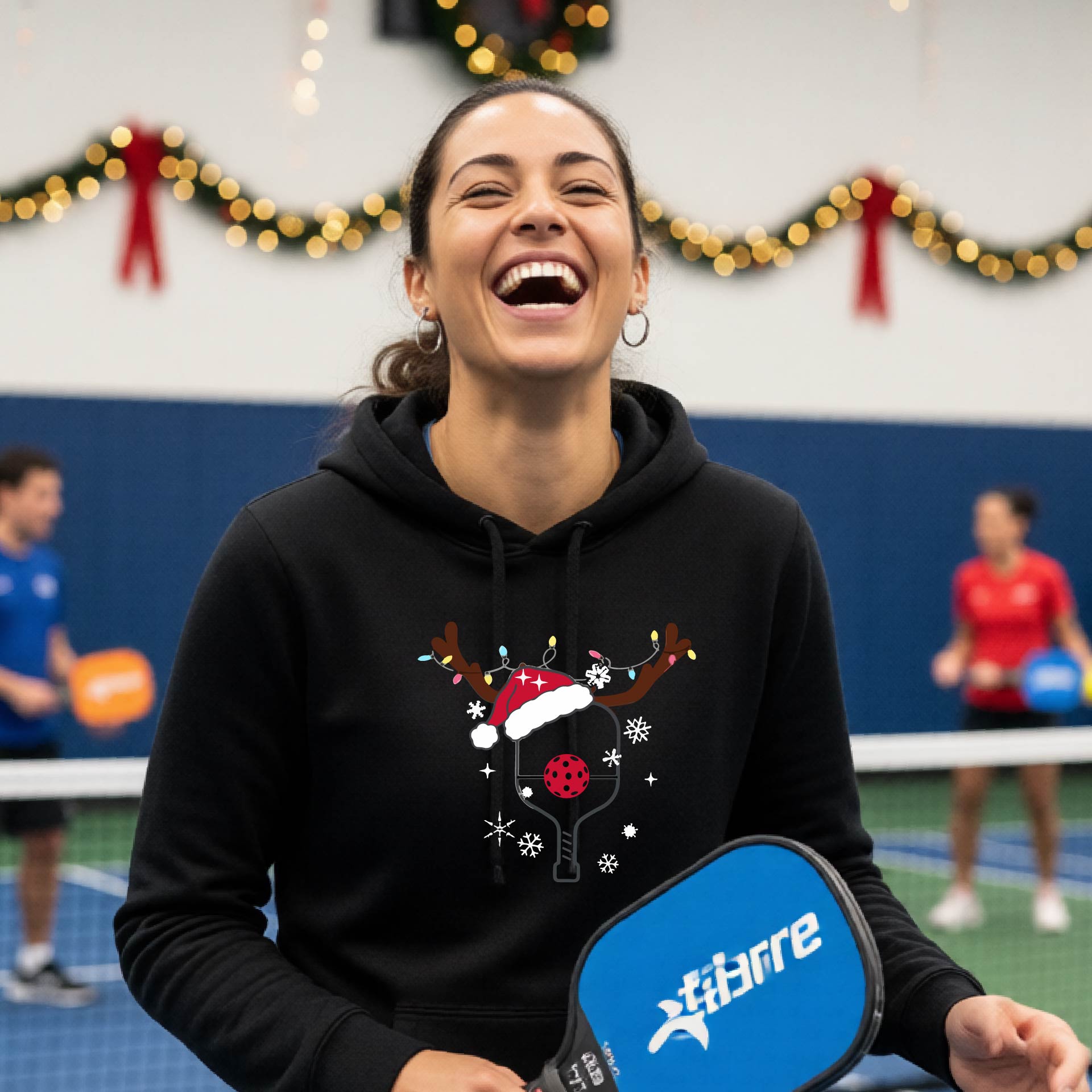 A black hooded sweatshirt featuring a pickleball graphic as pickleball paddle wearing a santa hat and antlers.  The backgro0und is Christmas themed. 