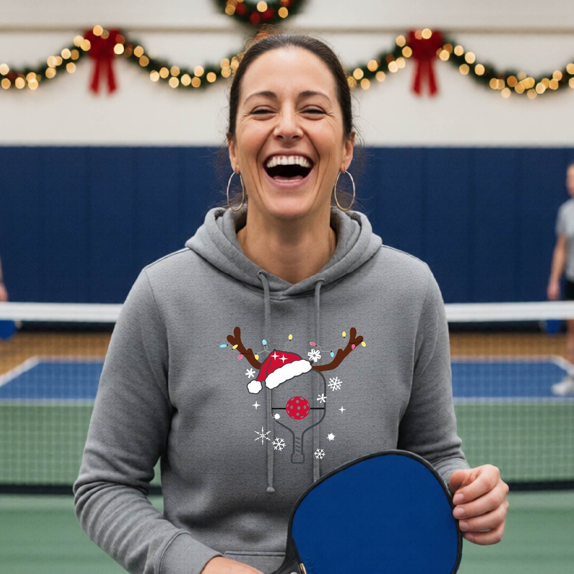 A gray hooded sweatshirt featuring a pickleball graphic as pickleball paddle wearing a santa hat and antlers.  The backgro0und is Christmas themed. 