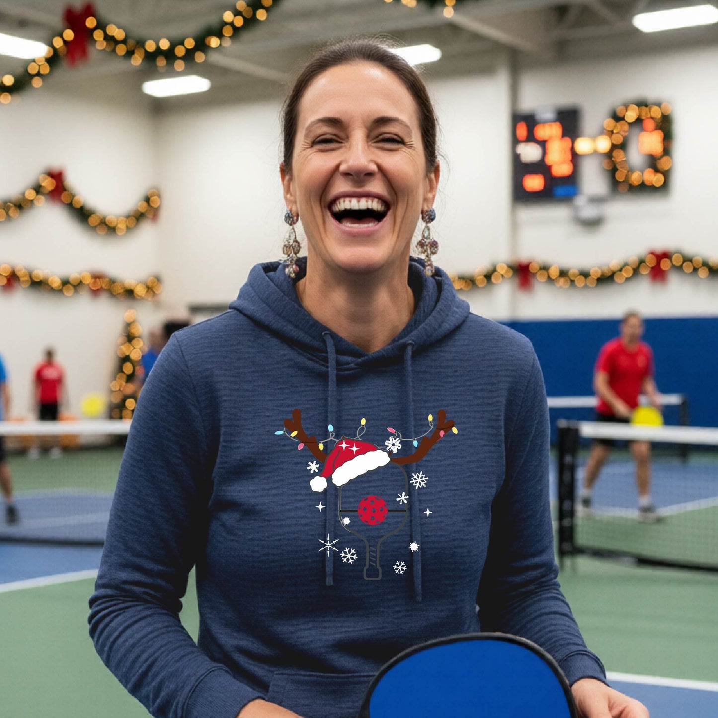 A navy hooded sweatshirt featuring a pickleball graphic as pickleball paddle wearing a santa hat and antlers.  The backgro0und is Christmas themed. 