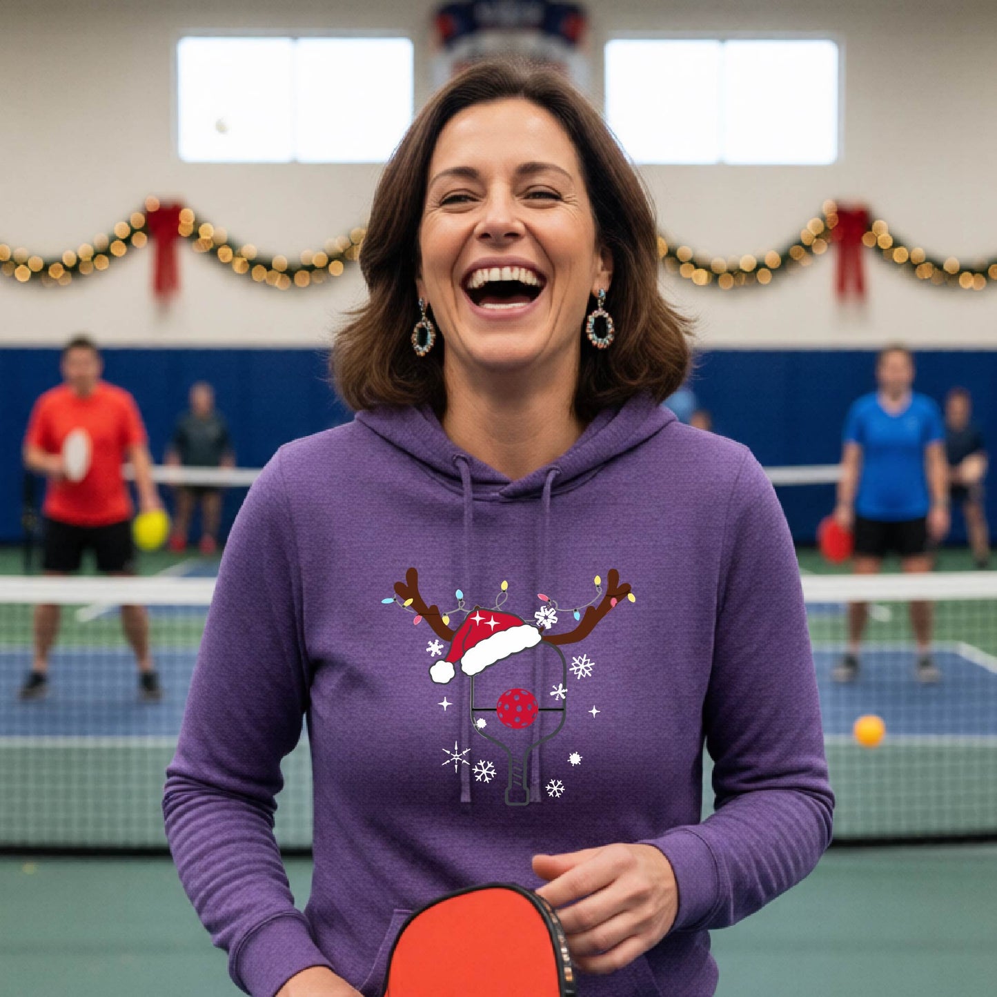 A purple hooded sweatshirt featuring a pickleball graphic as pickleball paddle wearing a santa hat and antlers.  The backgro0und is Christmas themed. 