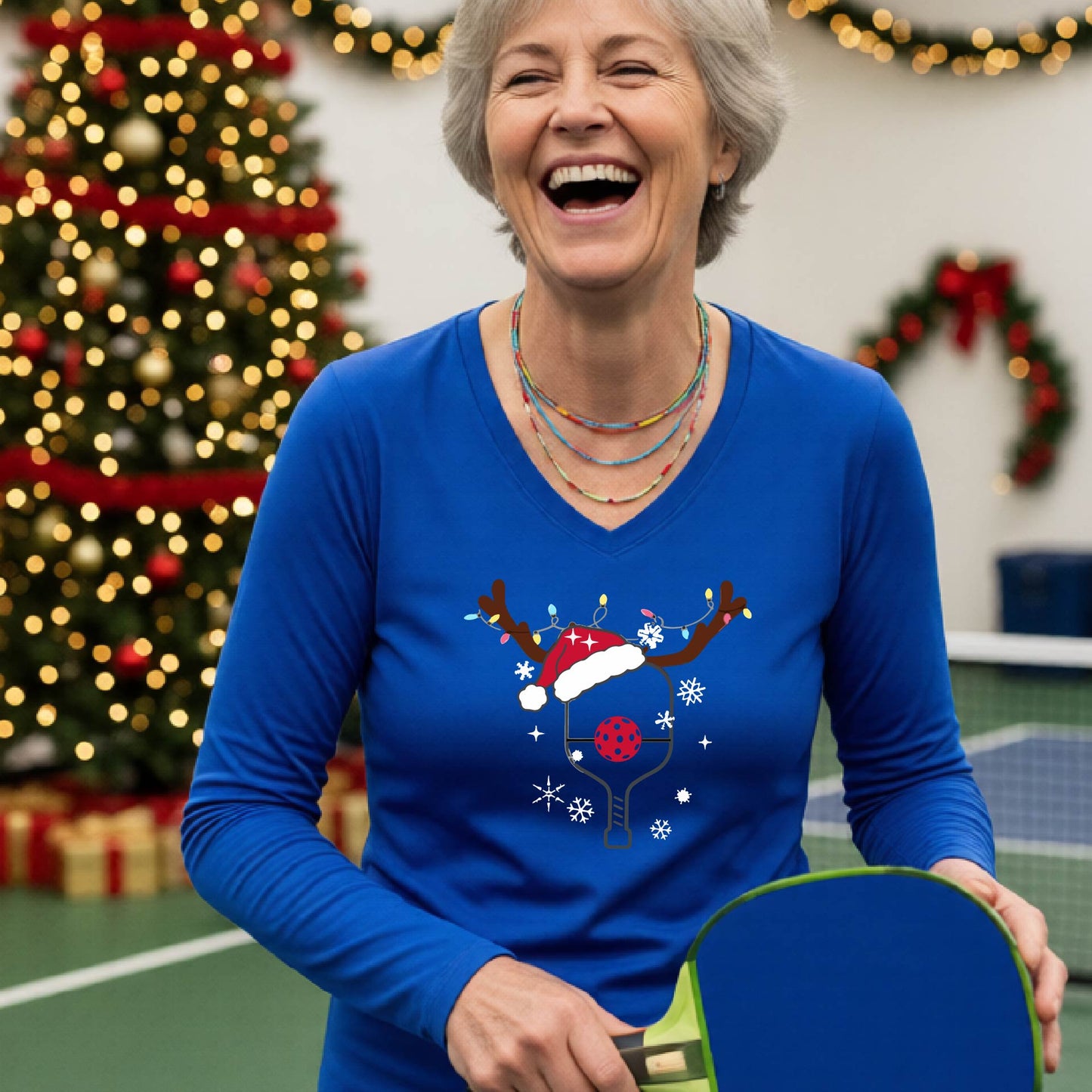 A woman laughing on a pickleball court wearing a royal blue long-sleeve V-neck shirt with a festive Christmas p pickleball paddle decorated with a Santa hat, reindeer antlers, and snowflakes.