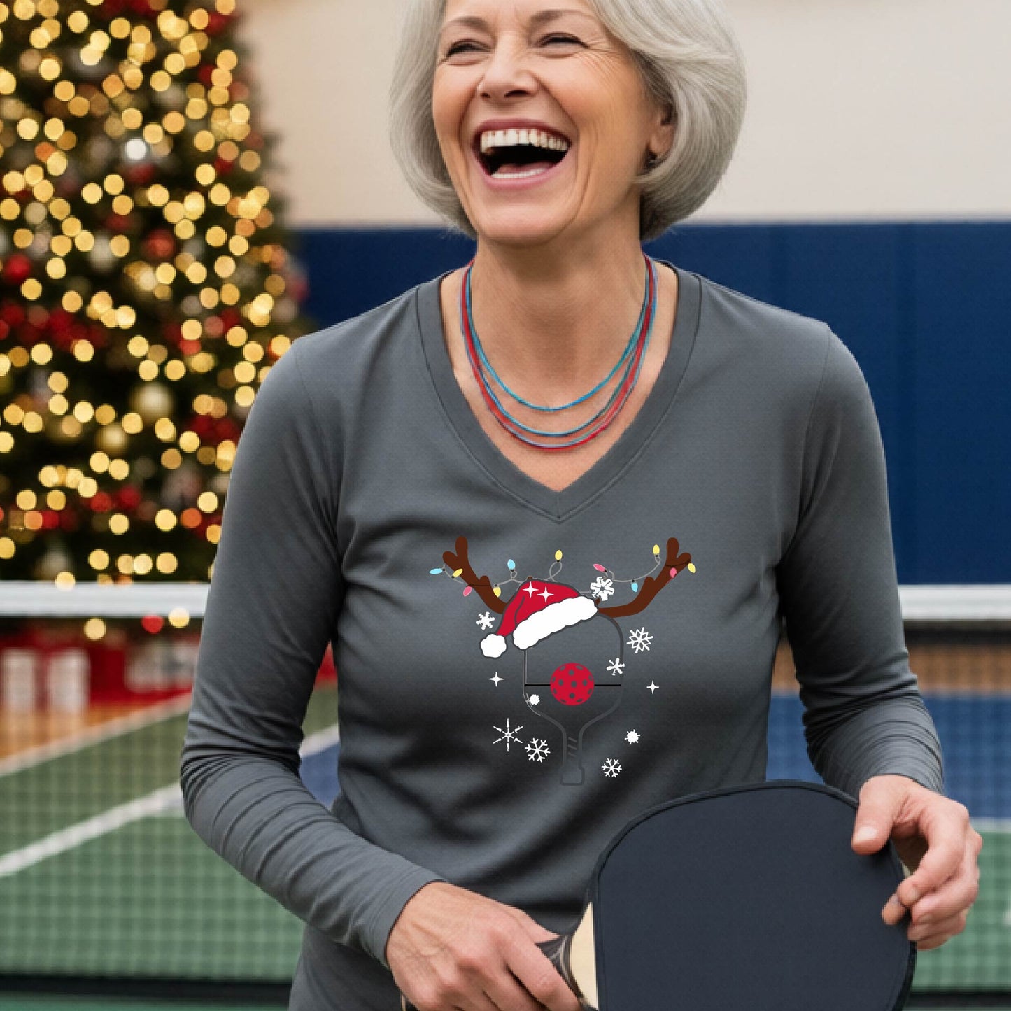 A woman laughing on a pickleball court wearing a gray long-sleeve V-neck shirt with a festive Christmas p pickleball paddle decorated with a Santa hat, reindeer antlers, and snowflakes.