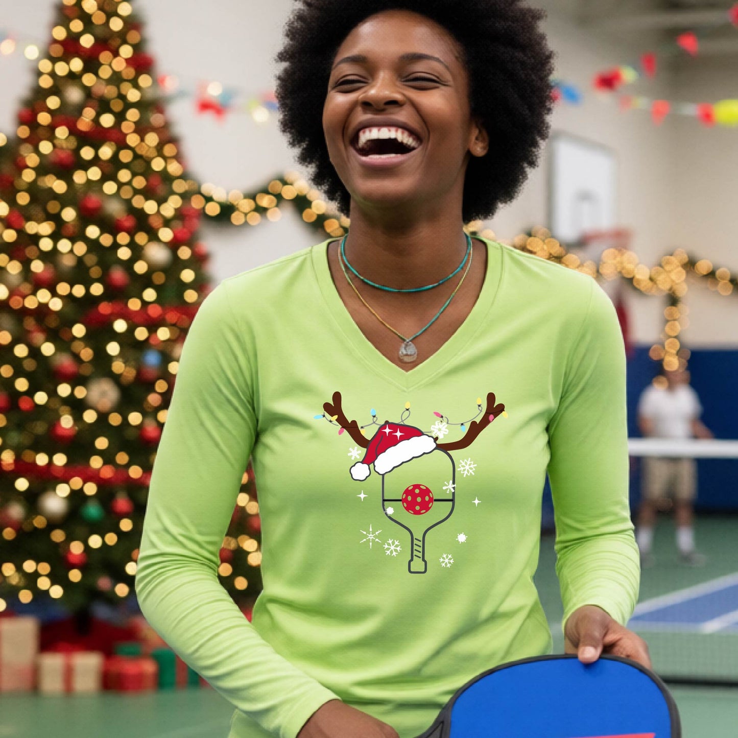 A woman laughing on a pickleball court wearing a lime green long-sleeve V-neck shirt with a festive Christmas p pickleball paddle decorated with a Santa hat, reindeer antlers, and snowflakes.