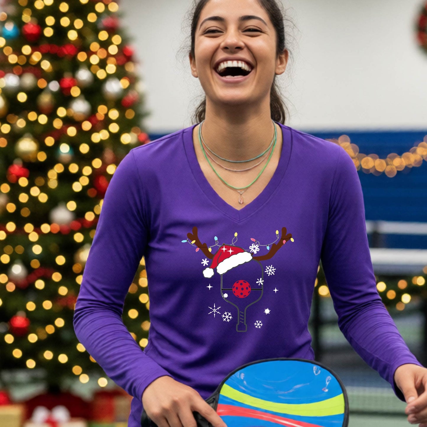 A woman laughing on a pickleball court wearing a purple long-sleeve V-neck shirt with a festive Christmas p pickleball paddle decorated with a Santa hat, reindeer antlers, and snowflakes.