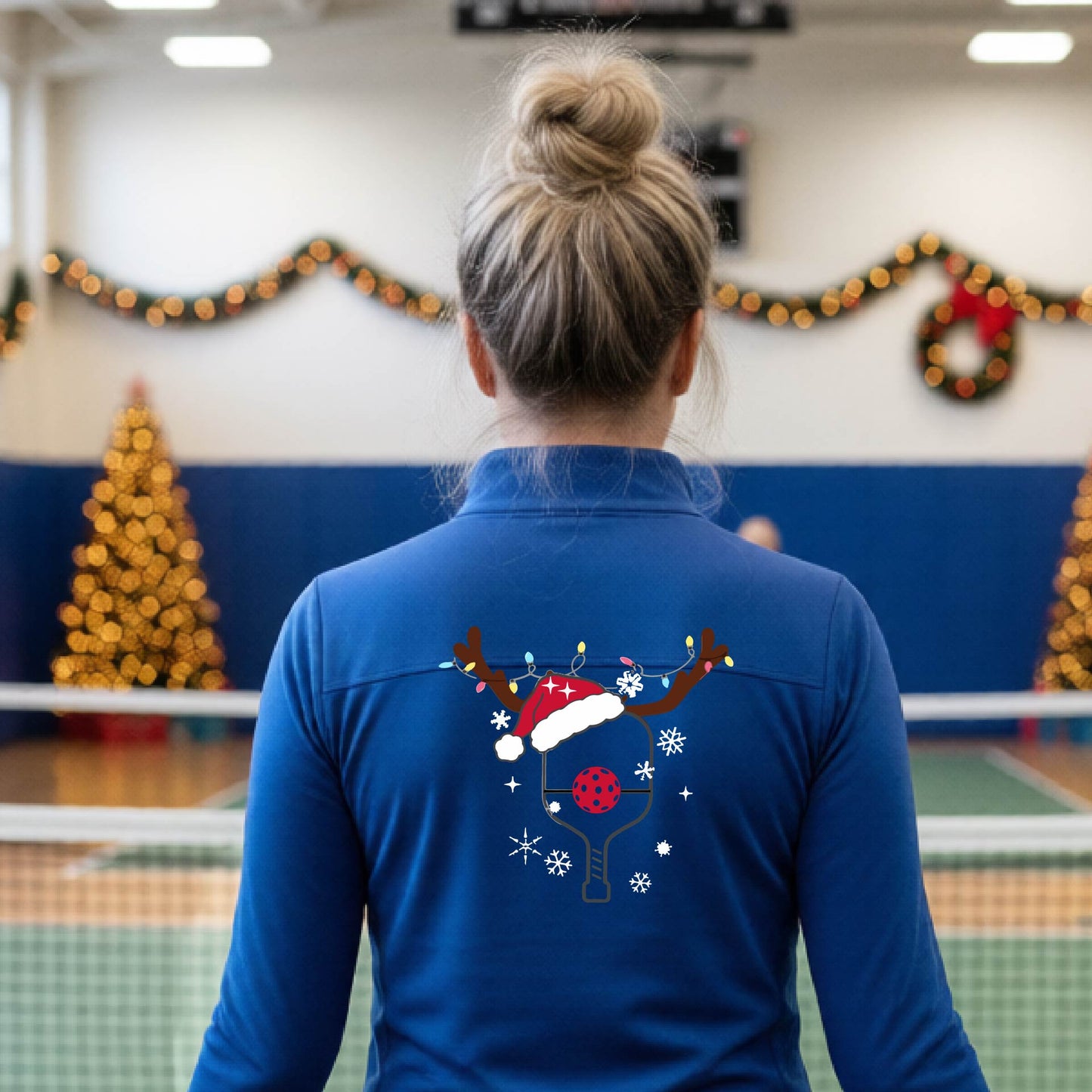 A woman viewed from behind wearing a royal blue long-sleeve athletic top with a Christmas-themed pickleball back graphic featuring reindeer antlers, a Santa hat, snowflakes, and a pickleball paddle. She is standing in a gymnasium with holiday decorations visible in the background.