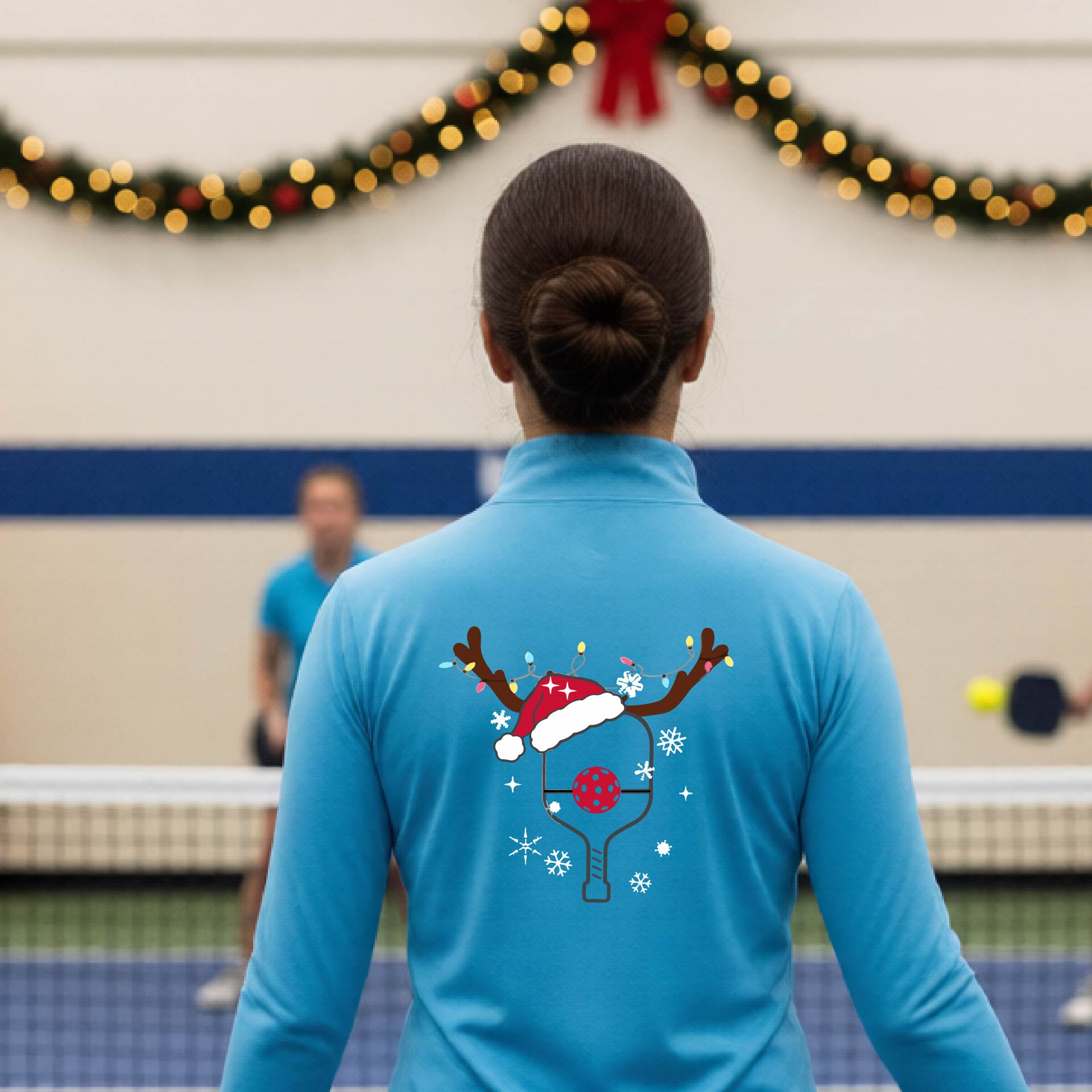 A woman viewed from behind wearing a light blue long-sleeve athletic top with a Christmas-themed pickleball back graphic featuring reindeer antlers, a Santa hat, snowflakes, and a pickleball paddle. She is standing in a gymnasium with holiday decorations visible in the background.