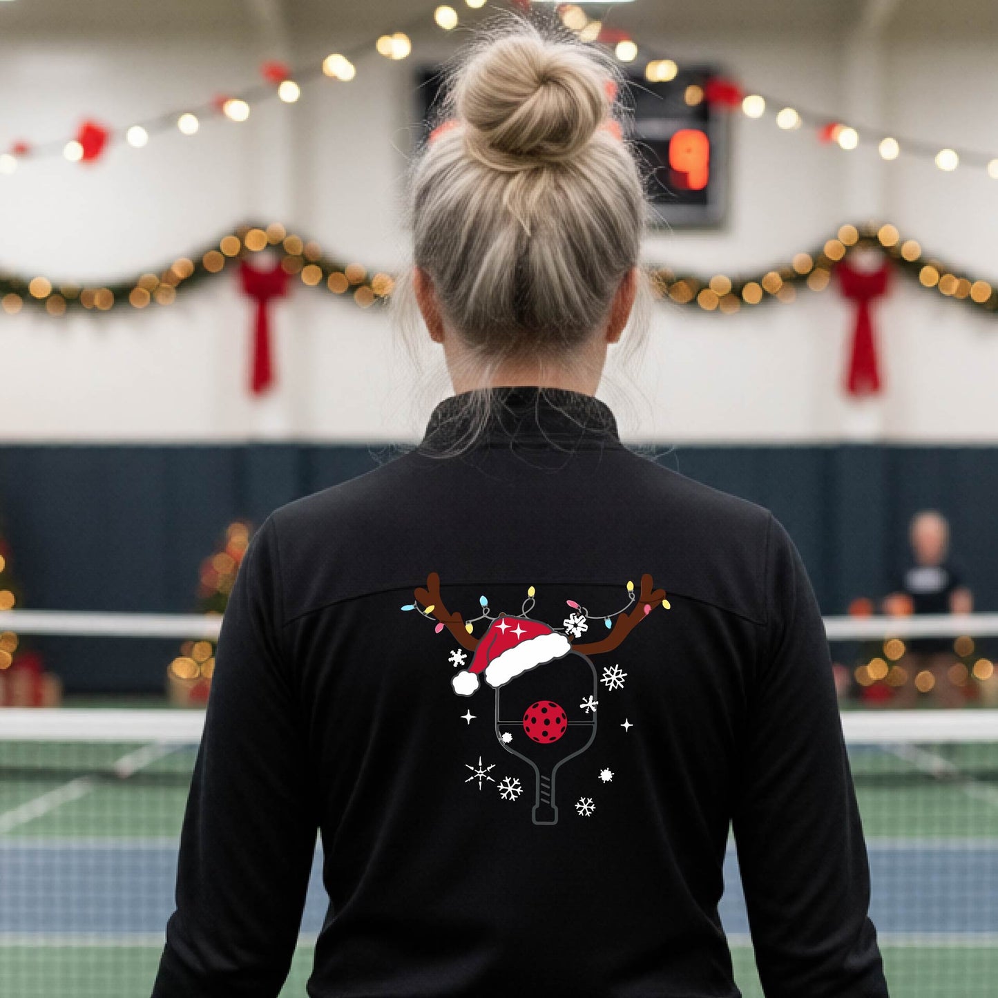 A woman viewed from behind wearing a black long-sleeve athletic top with a Christmas-themed pickleball back graphic featuring reindeer antlers, a Santa hat, snowflakes, and a pickleball paddle. She is standing in a gymnasium with holiday decorations visible in the background.