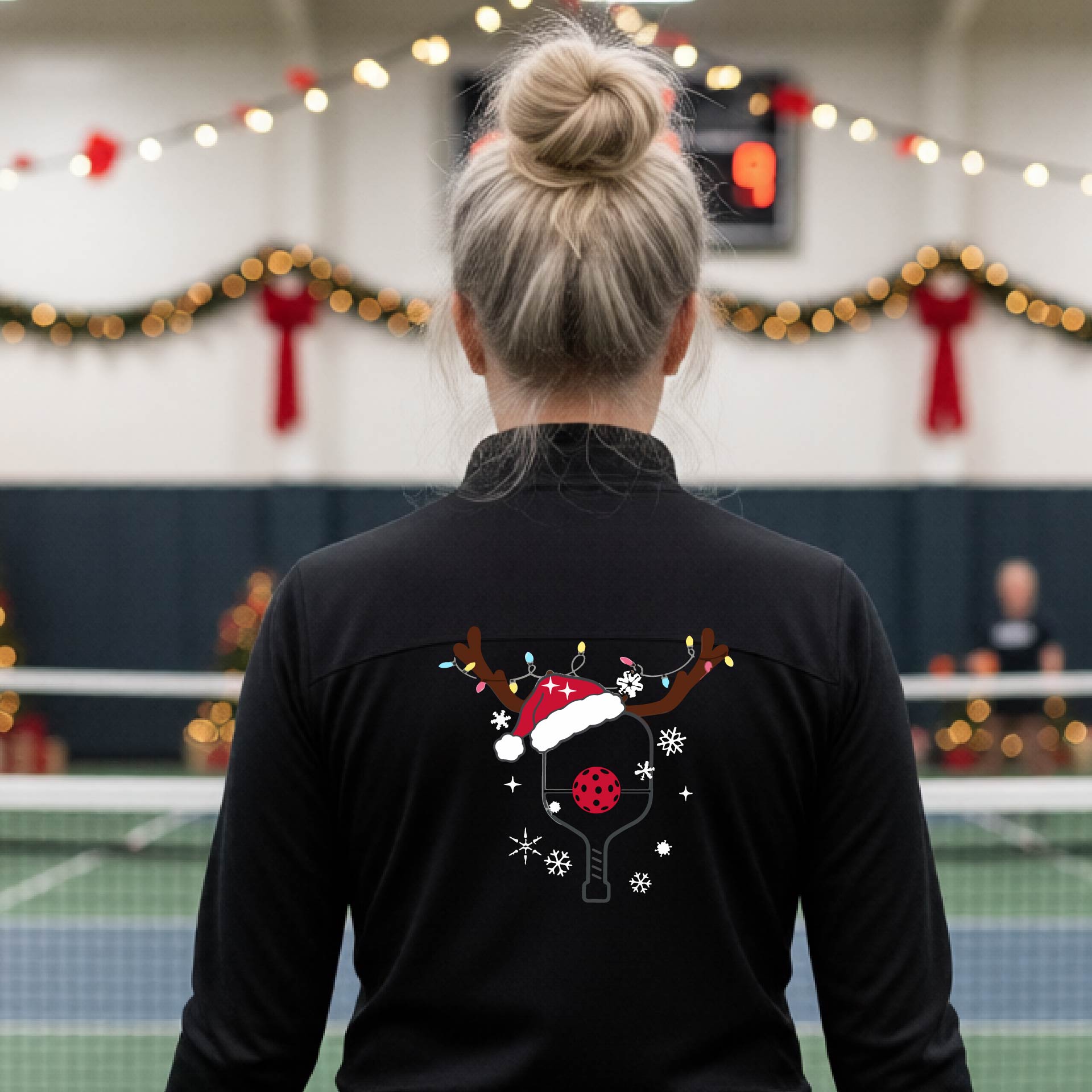 A woman viewed from behind wearing a black long-sleeve athletic top with a Christmas-themed pickleball back graphic featuring reindeer antlers, a Santa hat, snowflakes, and a pickleball paddle. She is standing in a gymnasium with holiday decorations visible in the background.