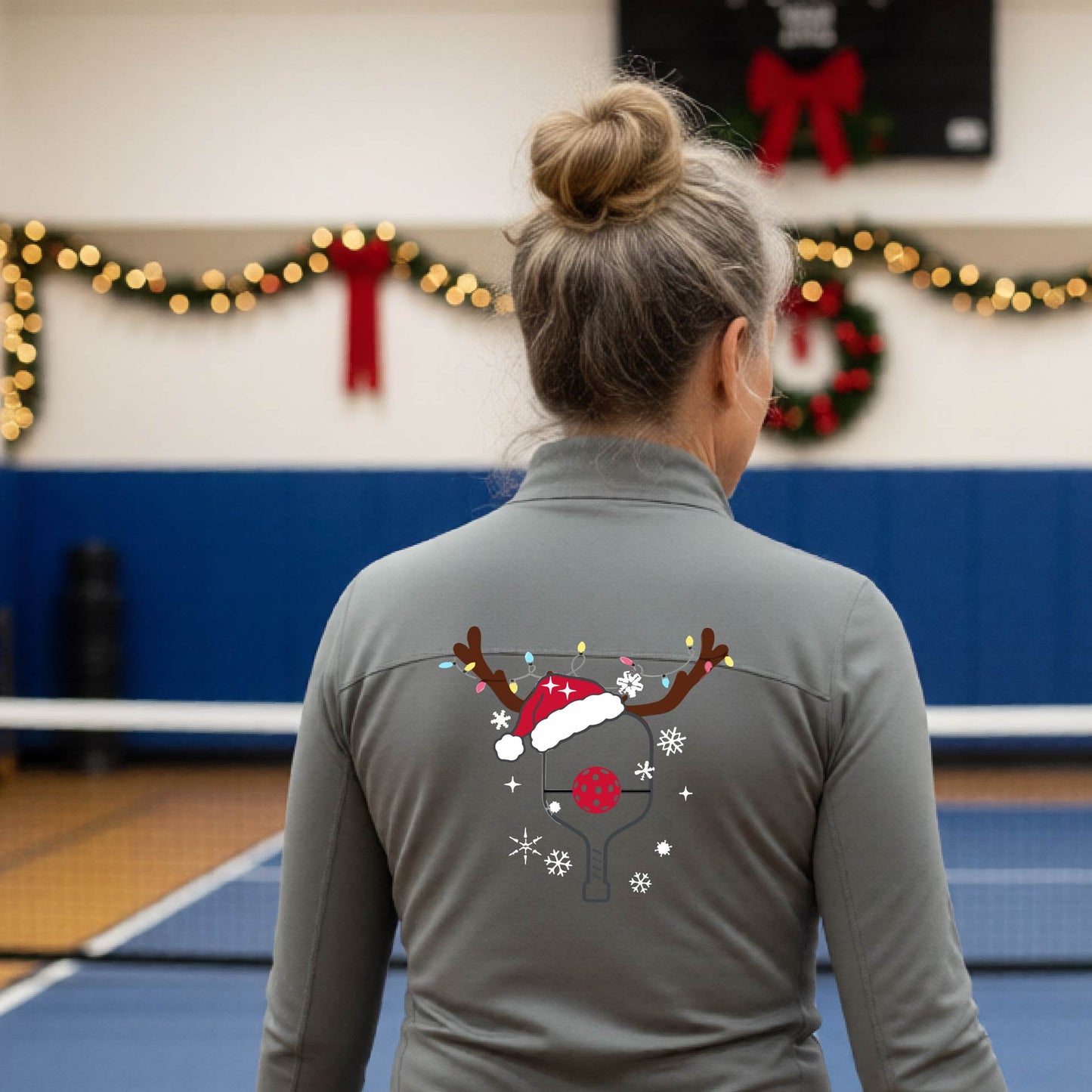 A woman viewed from behind wearing a gray long-sleeve athletic top with a Christmas-themed pickleball back graphic featuring reindeer antlers, a Santa hat, snowflakes, and a pickleball paddle. She is standing in a gymnasium with holiday decorations visible in the background.