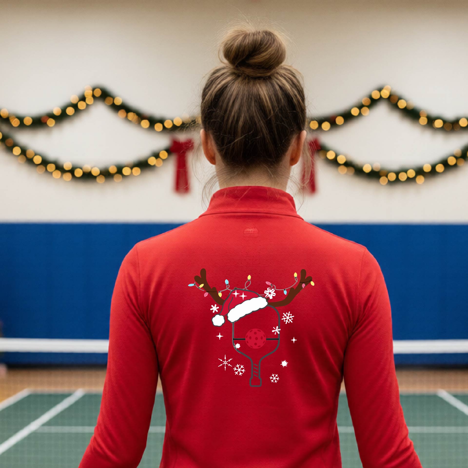 A woman viewed from behind wearing a red long-sleeve athletic top with a Christmas-themed pickleball back graphic featuring reindeer antlers, a Santa hat, snowflakes, and a pickleball paddle. She is standing in a gymnasium with holiday decorations visible in the background.