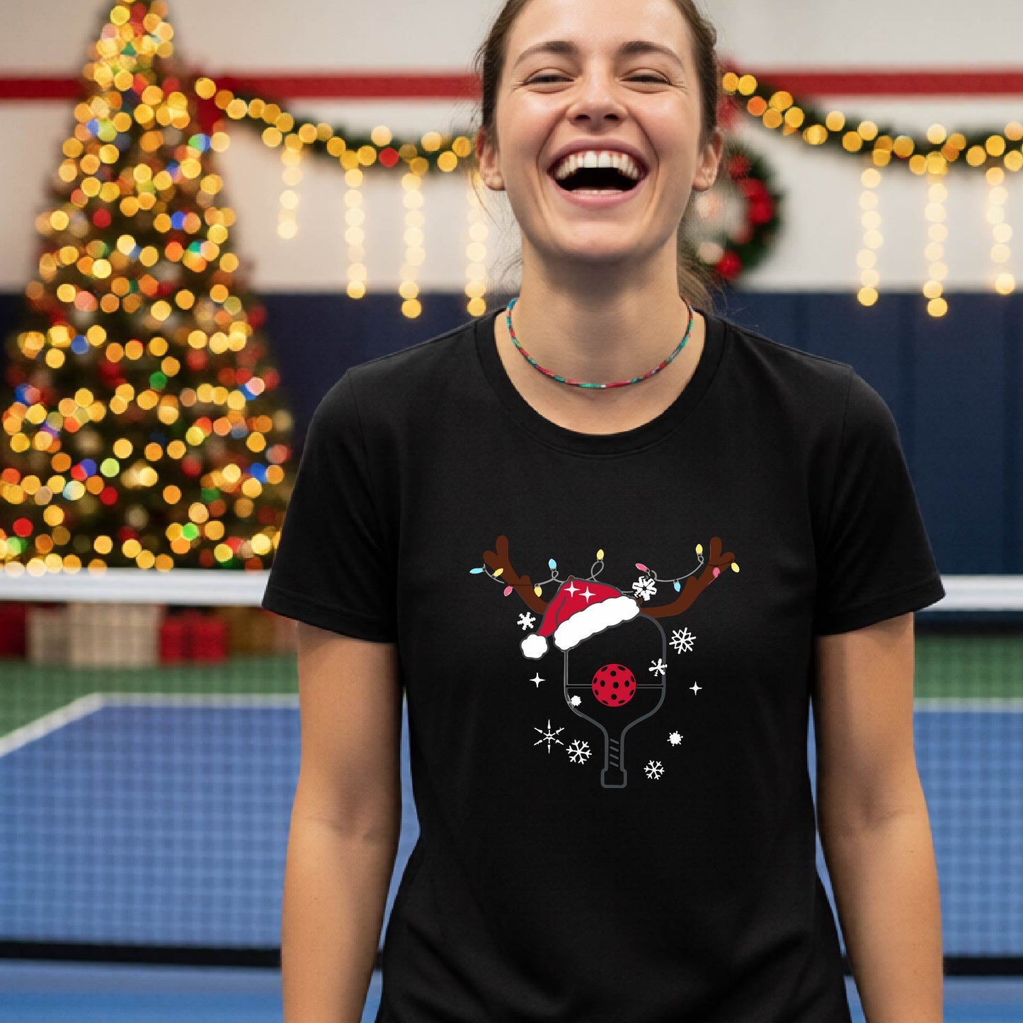 A woman wearing a black graphic t-shirt, standing on a pickleball court with Christmas decorations in the background. The t-shirt features a graphic of a pickleball paddle with reindeer antlers and a Santa hat.