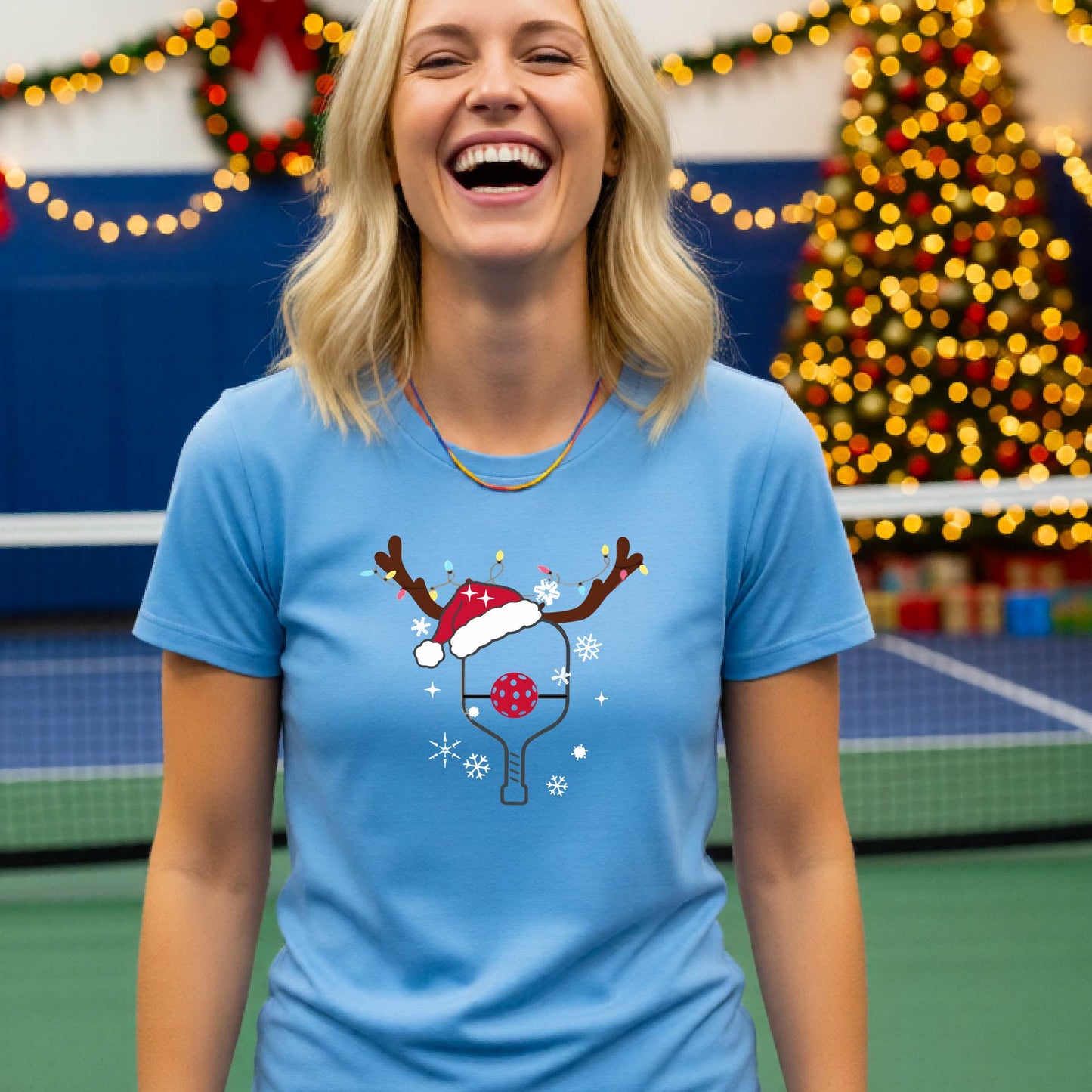 A woman wearing a light blue graphic t-shirt, standing on a pickleball court with Christmas decorations in the background. The t-shirt features a graphic of a pickleball paddle with reindeer antlers and a Santa hat.