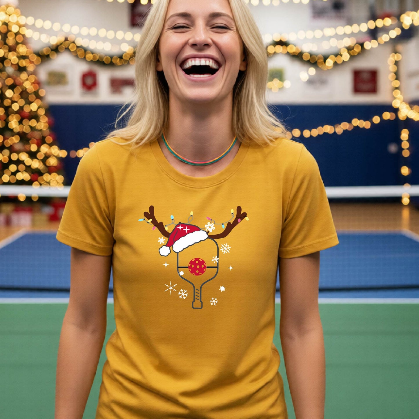 A woman wearing a mustard yellow gold graphic t-shirt, standing on a pickleball court with Christmas decorations in the background. The t-shirt features a graphic of a pickleball paddle with reindeer antlers and a Santa hat.
