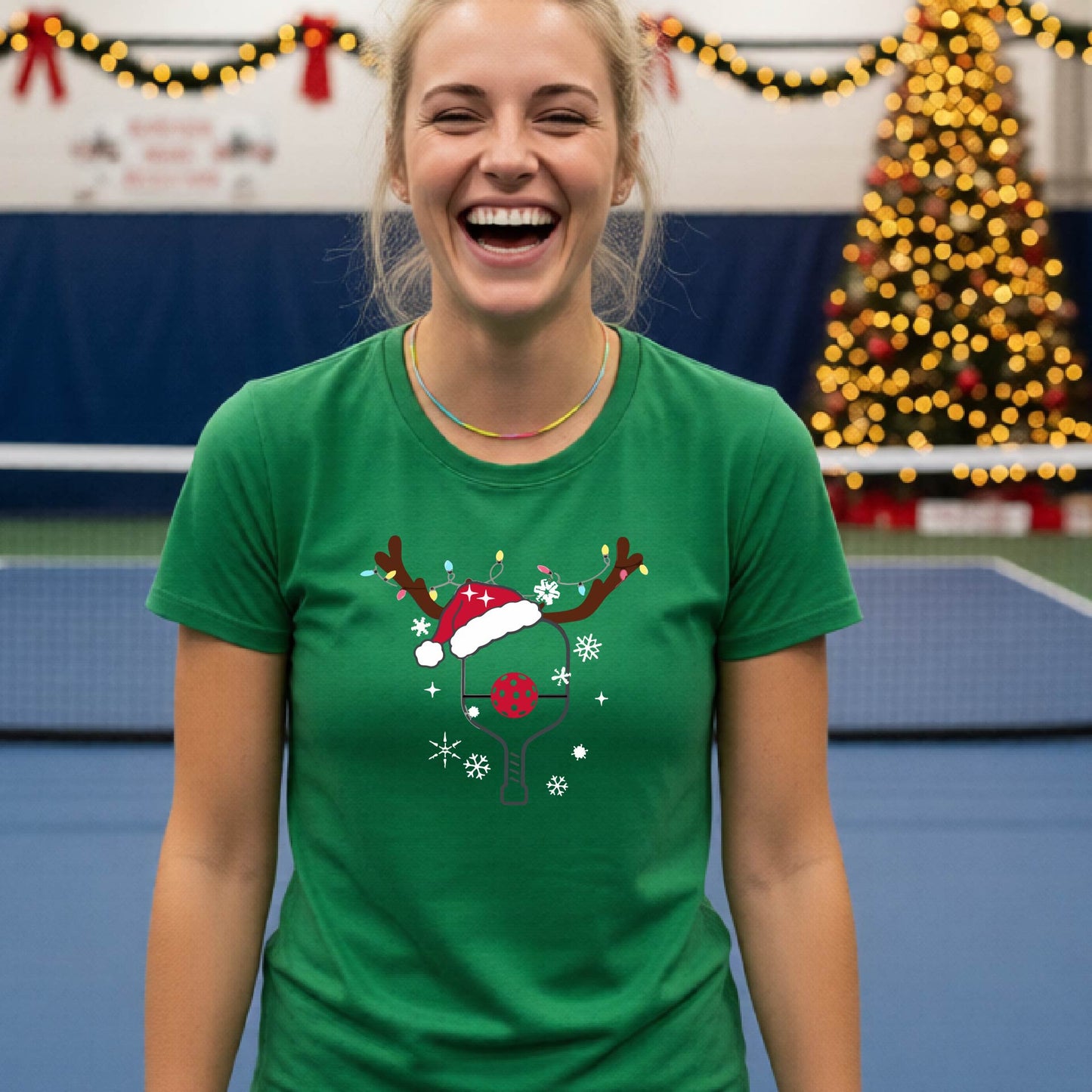 A woman wearing a green graphic t-shirt, standing on a pickleball court with Christmas decorations in the background. The t-shirt features a graphic of a pickleball paddle with reindeer antlers and a Santa hat.