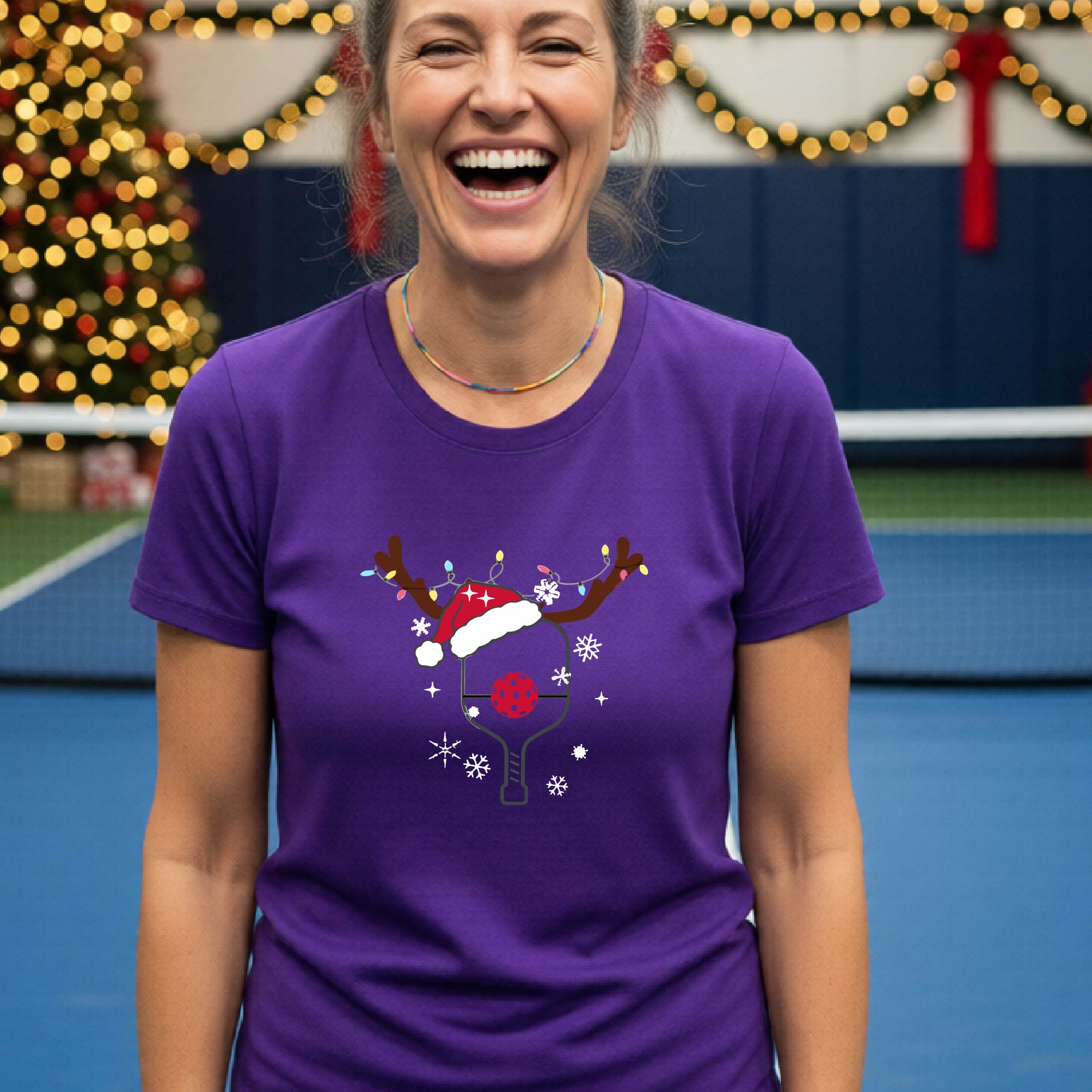 A woman wearing a bright purple graphic t-shirt, standing on a pickleball court with Christmas decorations in the background. The t-shirt features a graphic of a pickleball paddle with reindeer antlers and a Santa hat.