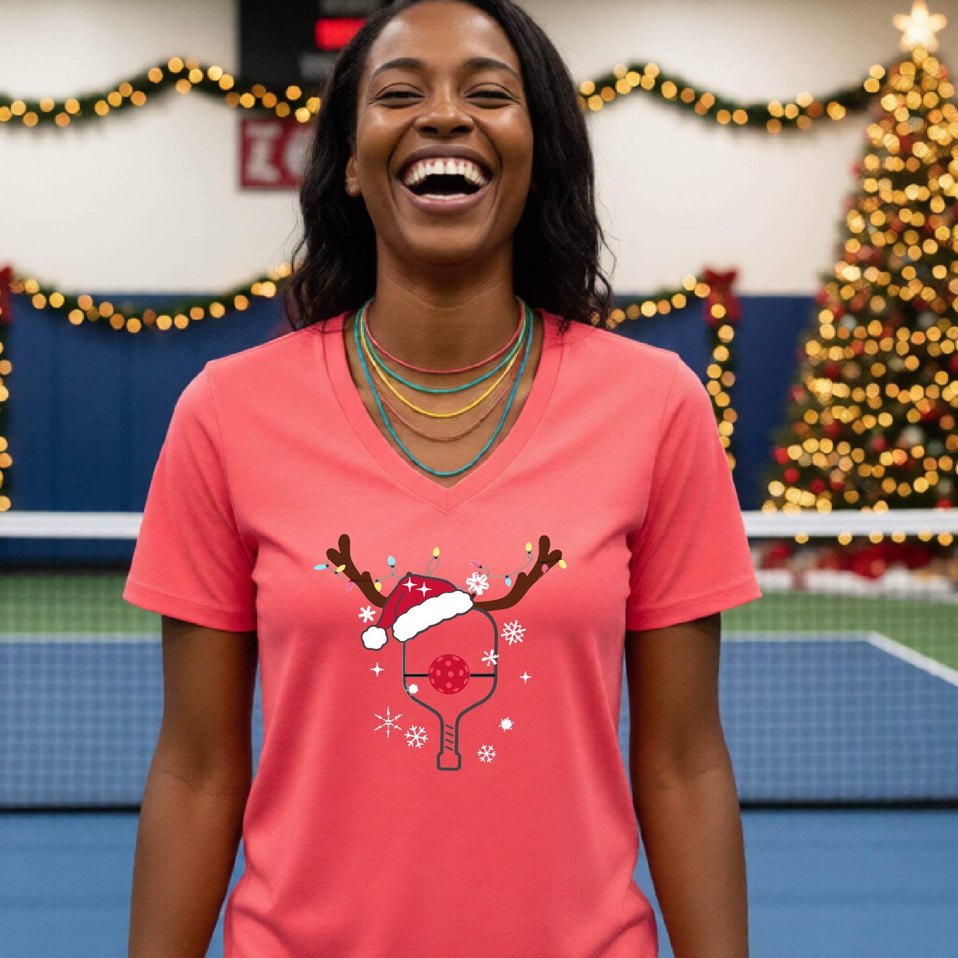 A woman is smiling and laughing while wearing a bright coral pink V-neck t-shirt with a Christmas-themed pickleball paddle with antlers and santa hat graphic with snowflakes, and a red pickleball nose. 