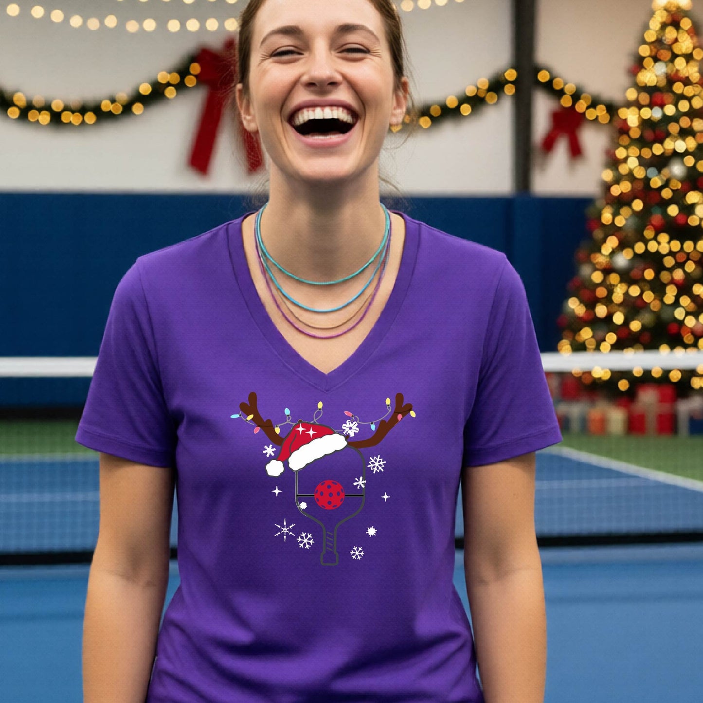 A woman is smiling and laughing while wearing a bright purple V-neck t-shirt with a Christmas-themed pickleball paddle with antlers and santa hat graphic with snowflakes, and a red pickleball nose. 