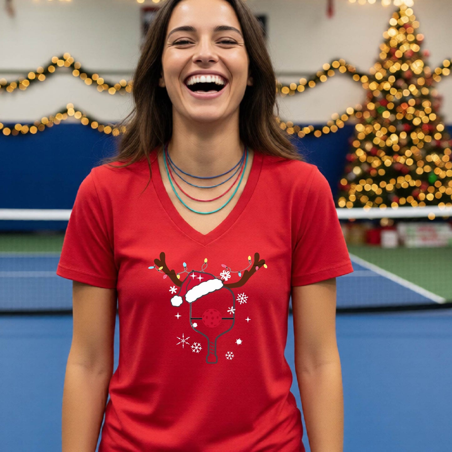 A woman is smiling and laughing while wearing a bright red V-neck t-shirt with a Christmas-themed pickleball paddle with antlers and santa hat graphic with snowflakes, and a red pickleball nose. 