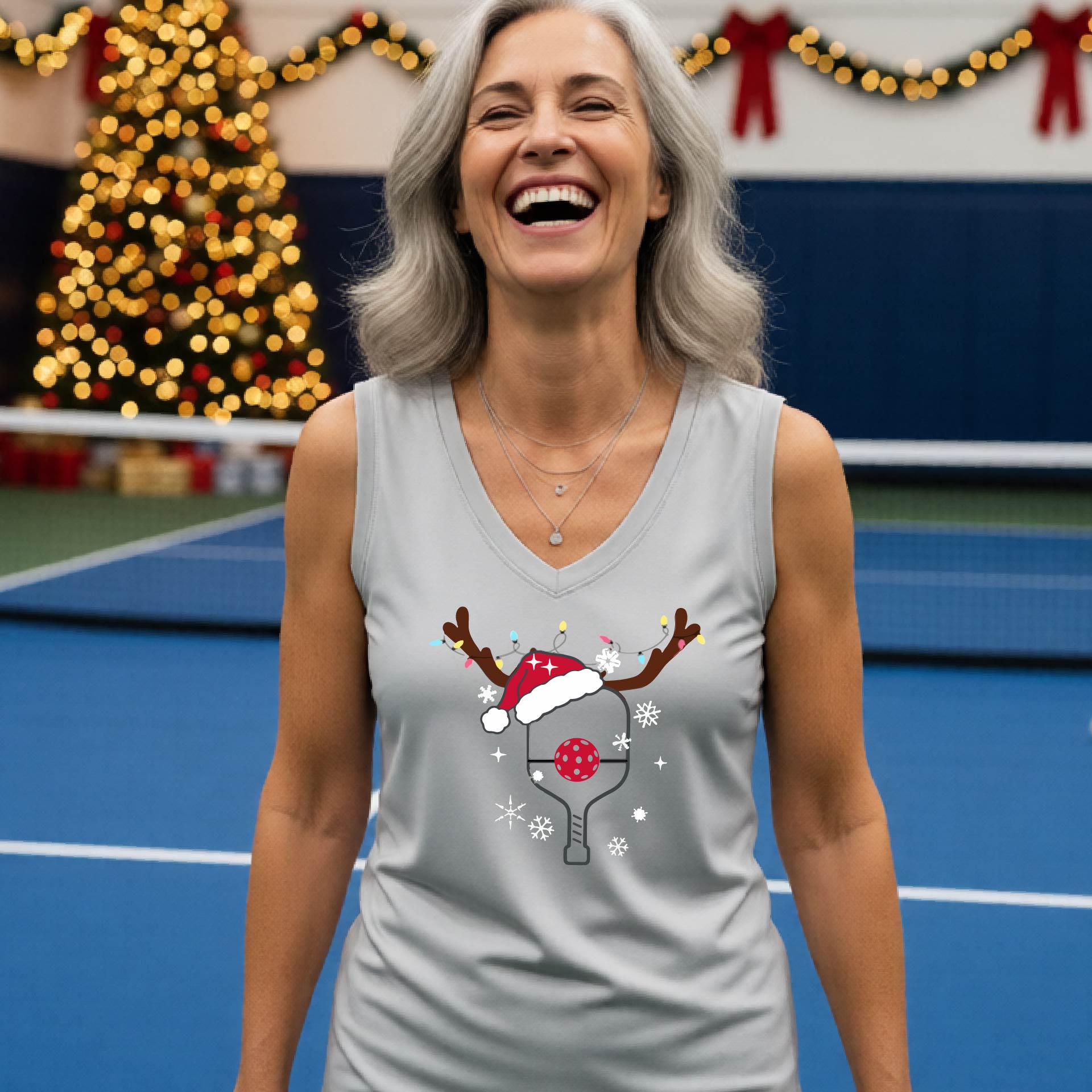 A smiling woman stands on a blue pickleball court wearing a light silver gray sleeveless tank top with a Christmas-themed pickleball graphic. The graphic features a picleball paddle decorated with a red Santa hat, a red ball, reindeer ears, and snowflakes.