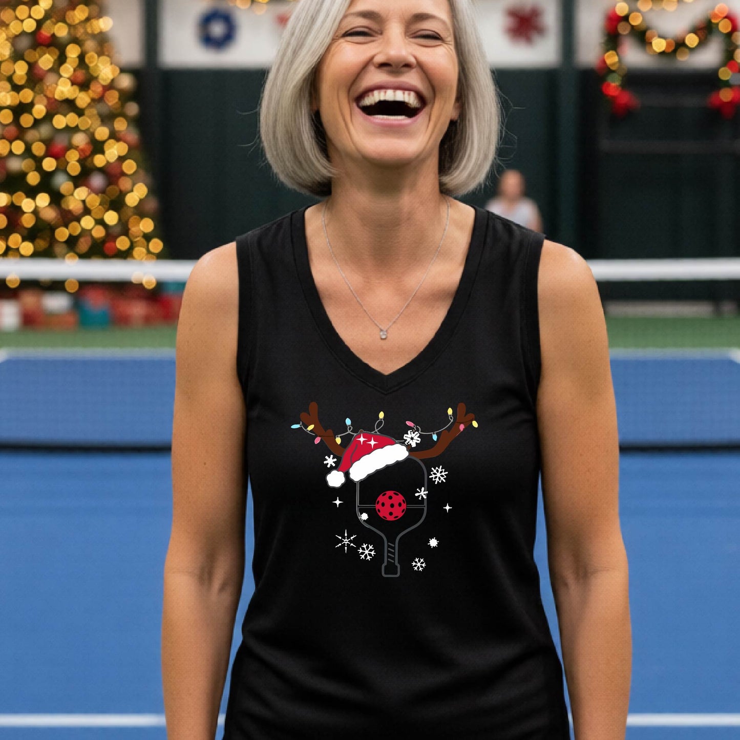 A smiling woman stands on a blue pickleball court wearing a black sleeveless tank top with a Christmas-themed pickleball graphic. The graphic features a picleball paddle decorated with a red Santa hat, a red ball, reindeer ears, and snowflakes.