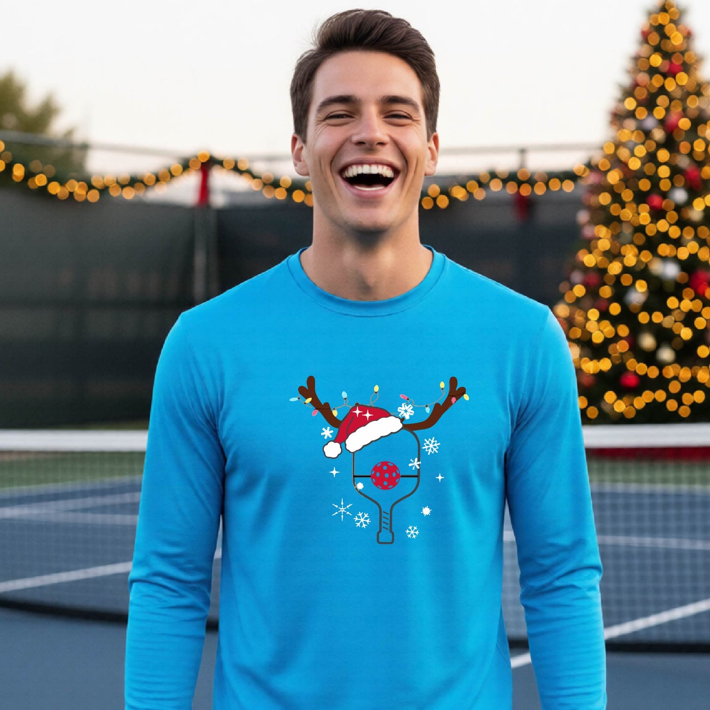 A man wearing a bright blue graphic t-shirt with a Christmas-themed pickleball design featuring a pickleball paddle wearing a Santa hat and reindeer antlers, and snowflakes, set against a blurred background of a pickleball court and holiday lights.