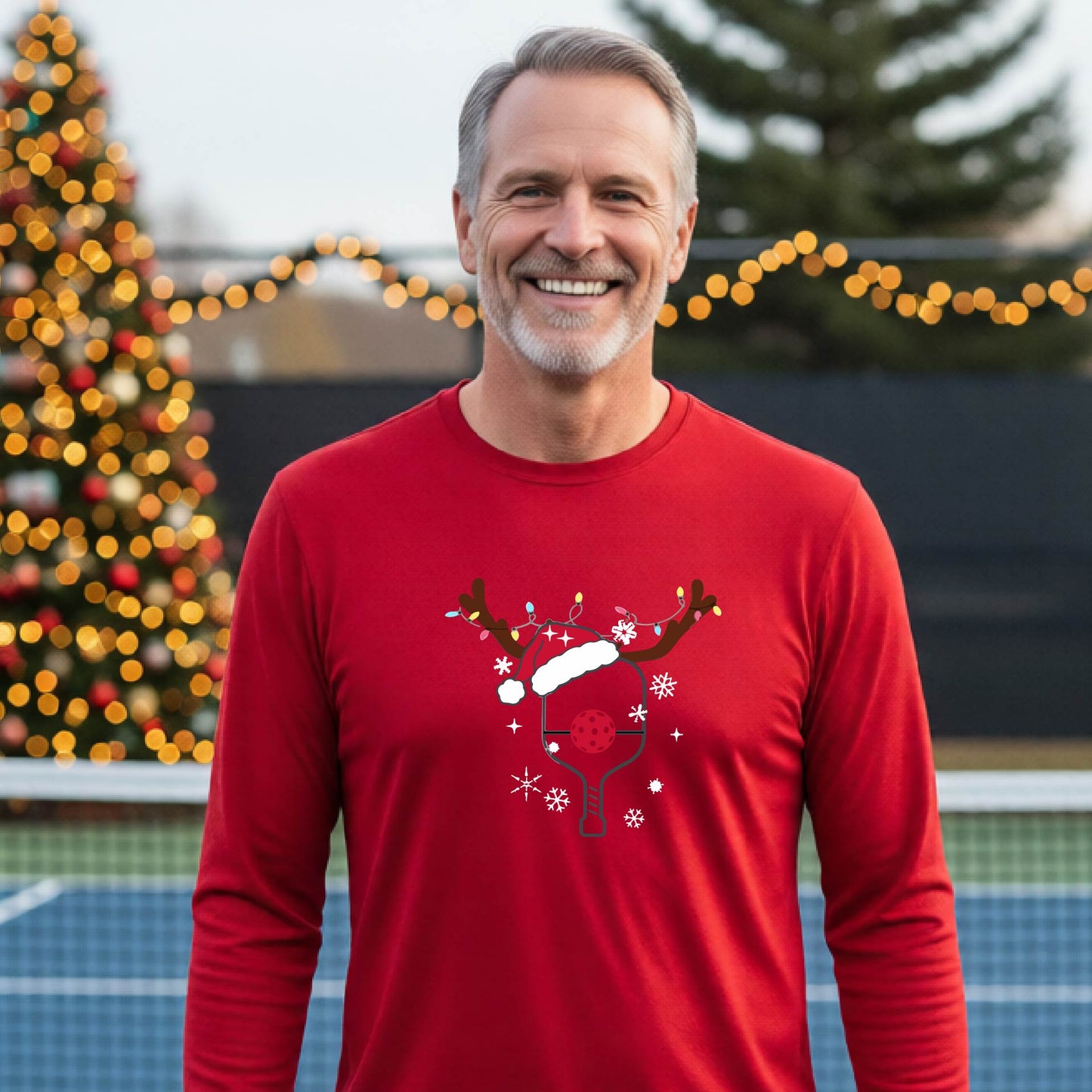 A man wearing a red graphic t-shirt with a Christmas-themed pickleball design featuring a pickleball paddle wearing a Santa hat and reindeer antlers, and snowflakes, set against a blurred background of a pickleball court and holiday lights.