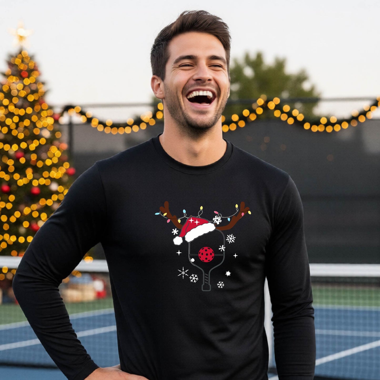 A man wearing a black graphic t-shirt with a Christmas-themed pickleball design featuring a pickleball paddle wearing a Santa hat and reindeer antlers, and snowflakes, set against a blurred background of a pickleball court and holiday lights.
