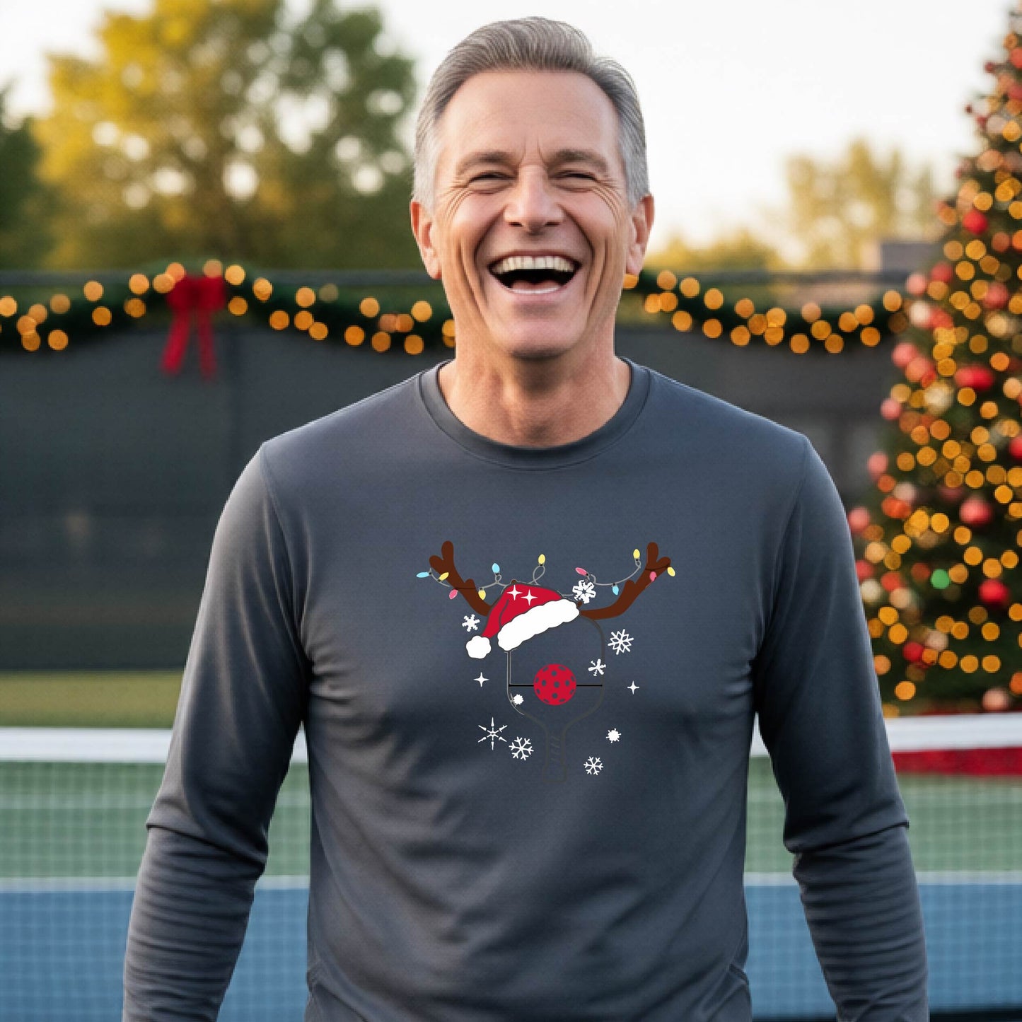 A man wearing a gray graphic t-shirt with a Christmas-themed pickleball design featuring a pickleball paddle wearing a Santa hat and reindeer antlers, and snowflakes, set against a blurred background of a pickleball court and holiday lights.