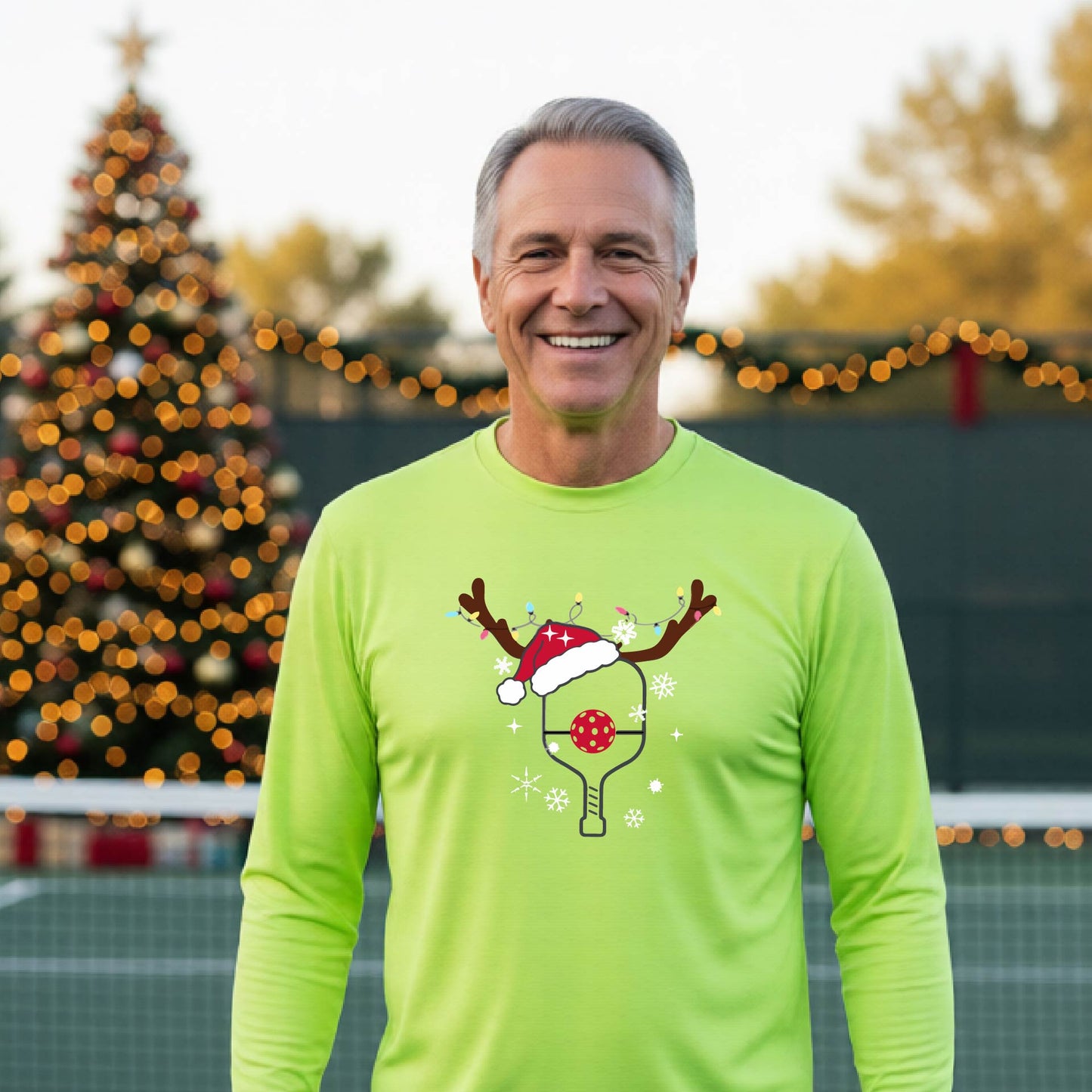 A man wearing a lime green graphic t-shirt with a Christmas-themed pickleball design featuring a pickleball paddle wearing a Santa hat and reindeer antlers, and snowflakes, set against a blurred background of a pickleball court and holiday lights.