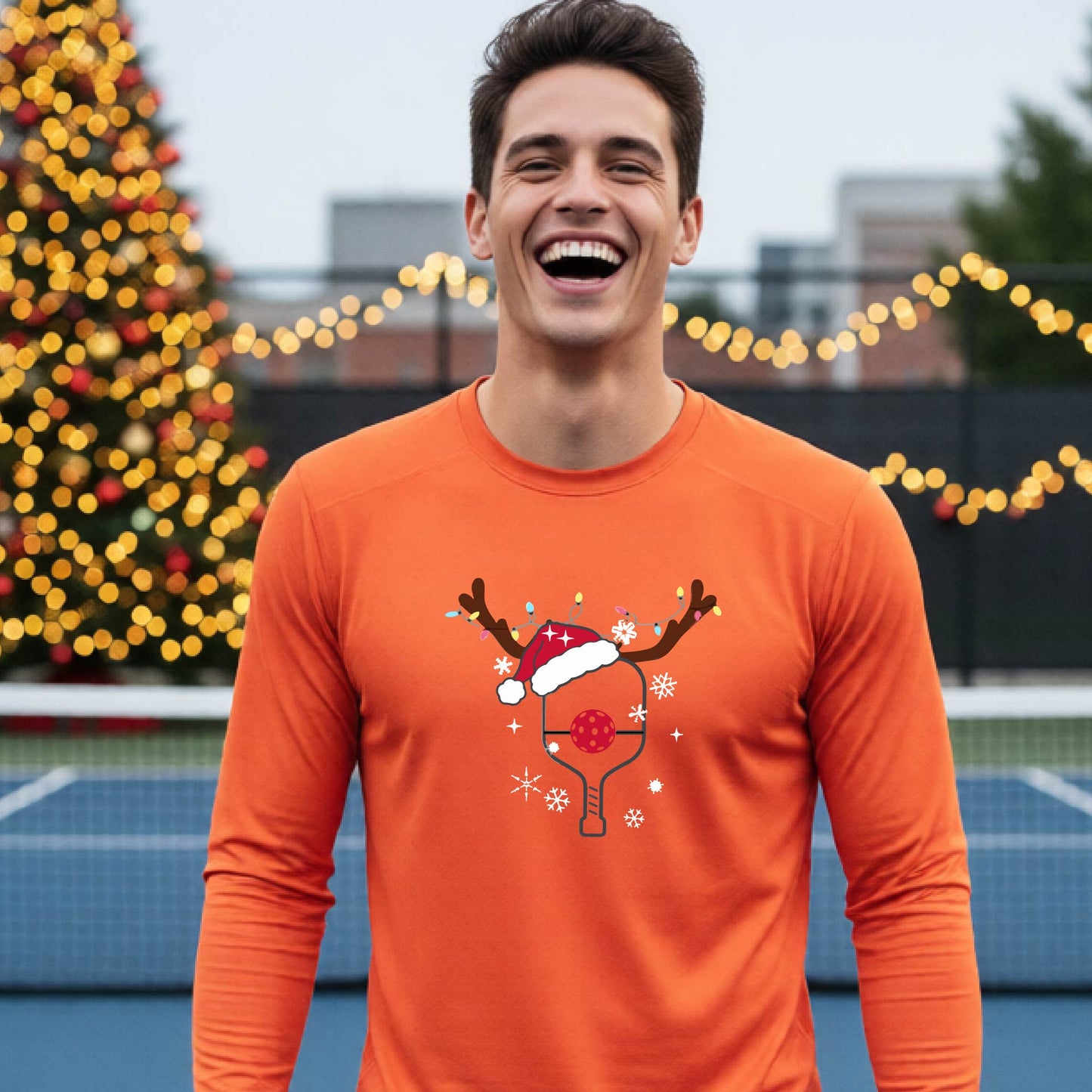 A man wearing a bright orange graphic t-shirt with a Christmas-themed pickleball design featuring a pickleball paddle wearing a Santa hat and reindeer antlers, and snowflakes, set against a blurred background of a pickleball court and holiday lights.