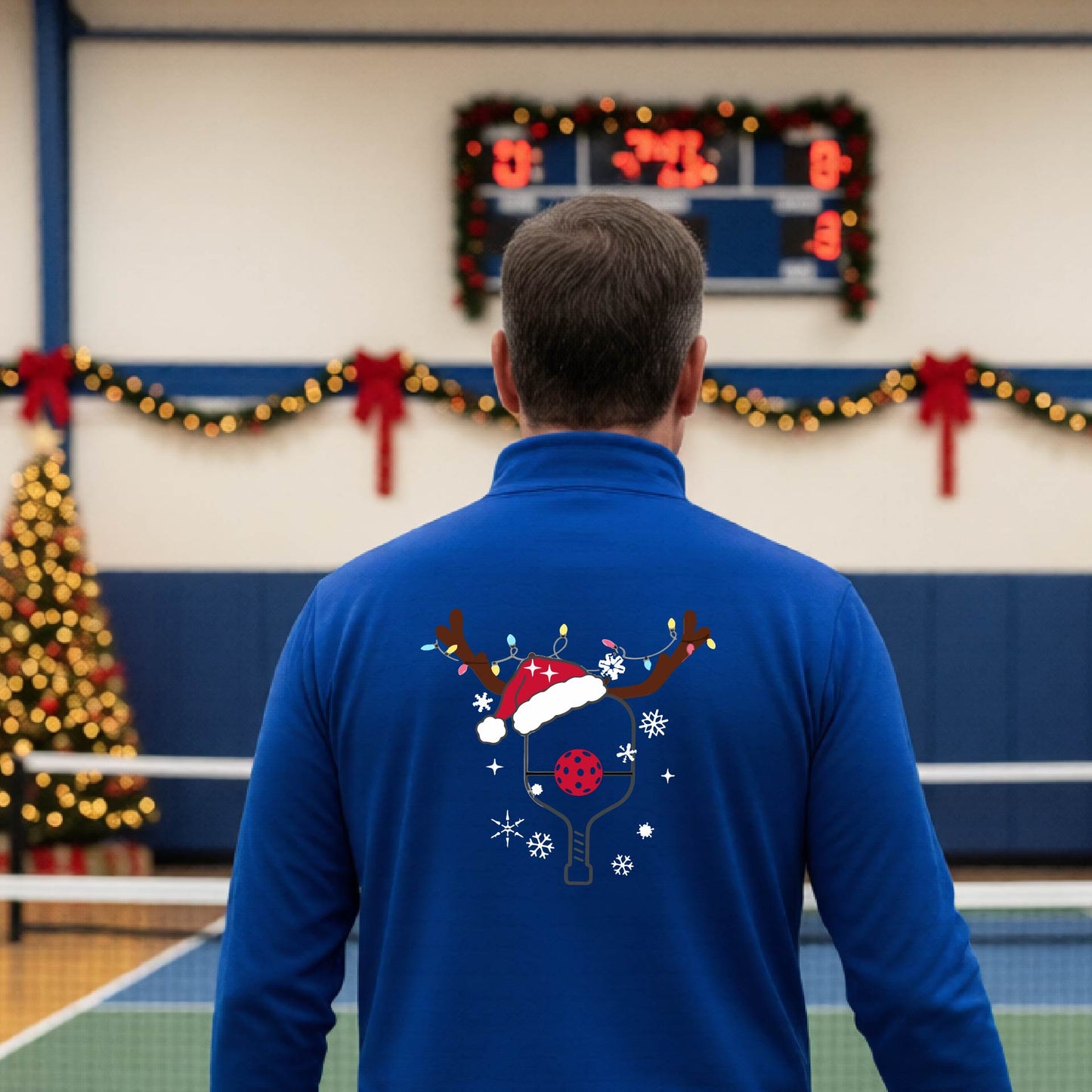 A man viewed from behind wearing a royal blue long-sleeve quarter-zip athletic shirt with a Christmas-themed back graphic of a pickleball paddle under a reindeer antler Santa hat. He is standing onpickleball court with holiday decorations.