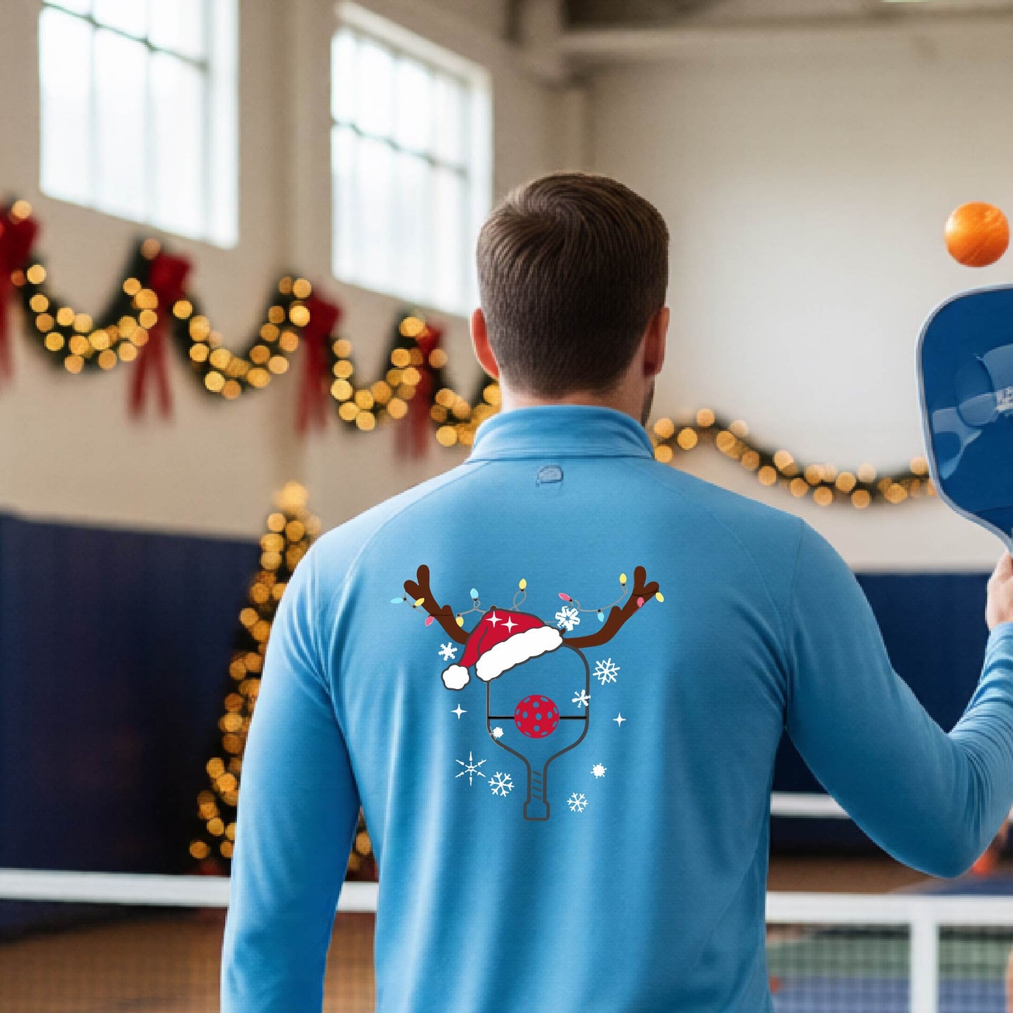 A man viewed from behind wearing a light blue long-sleeve quarter-zip athletic shirt with a Christmas-themed back graphic of a pickleball paddle under a reindeer antler Santa hat. He is standing onpickleball court with holiday decorations.