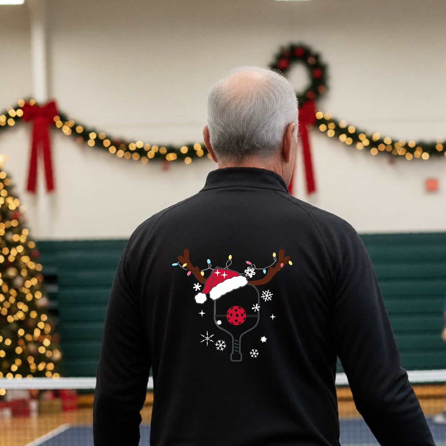 A man viewed from behind wearing a black long-sleeve quarter-zip athletic shirt with a Christmas-themed back graphic of a pickleball paddle under a reindeer antler Santa hat. He is standing onpickleball court with holiday decorations.