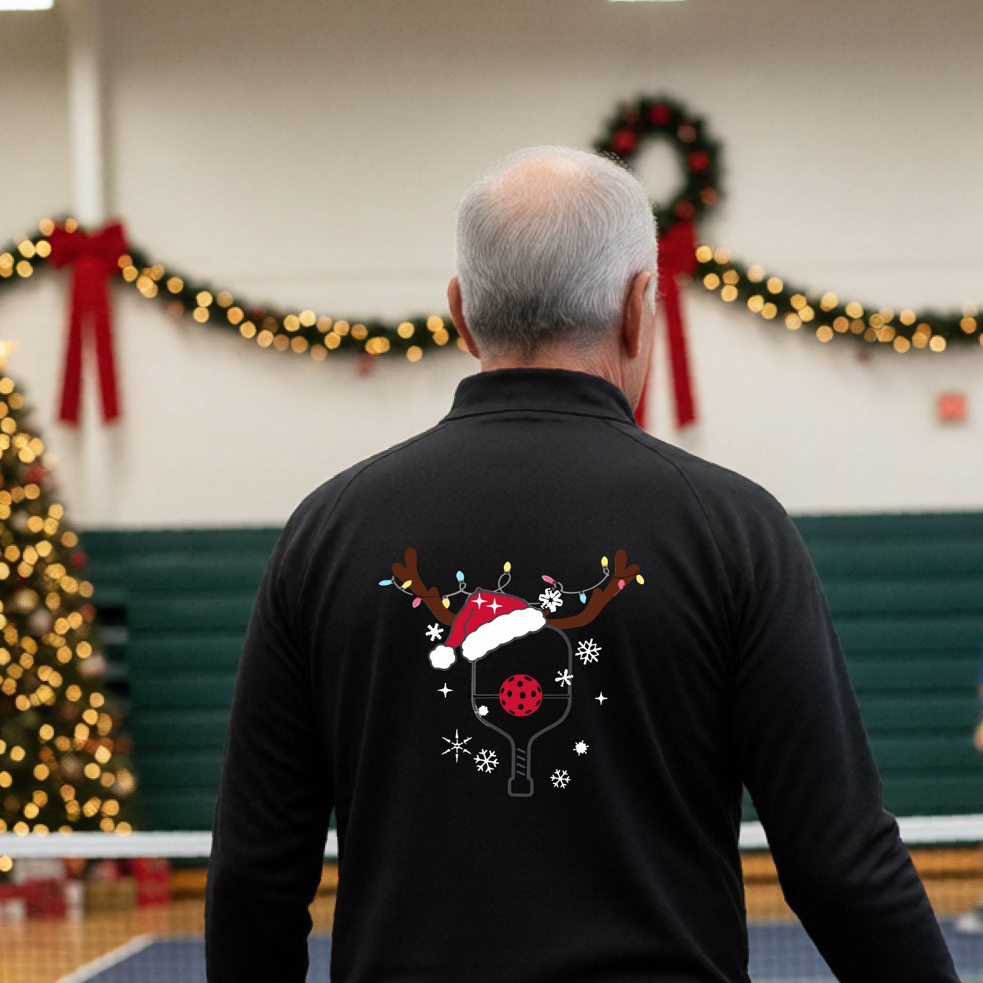 A man viewed from behind wearing a black long-sleeve quarter-zip athletic shirt with a Christmas-themed back graphic of a pickleball paddle under a reindeer antler Santa hat. He is standing onpickleball court with holiday decorations.