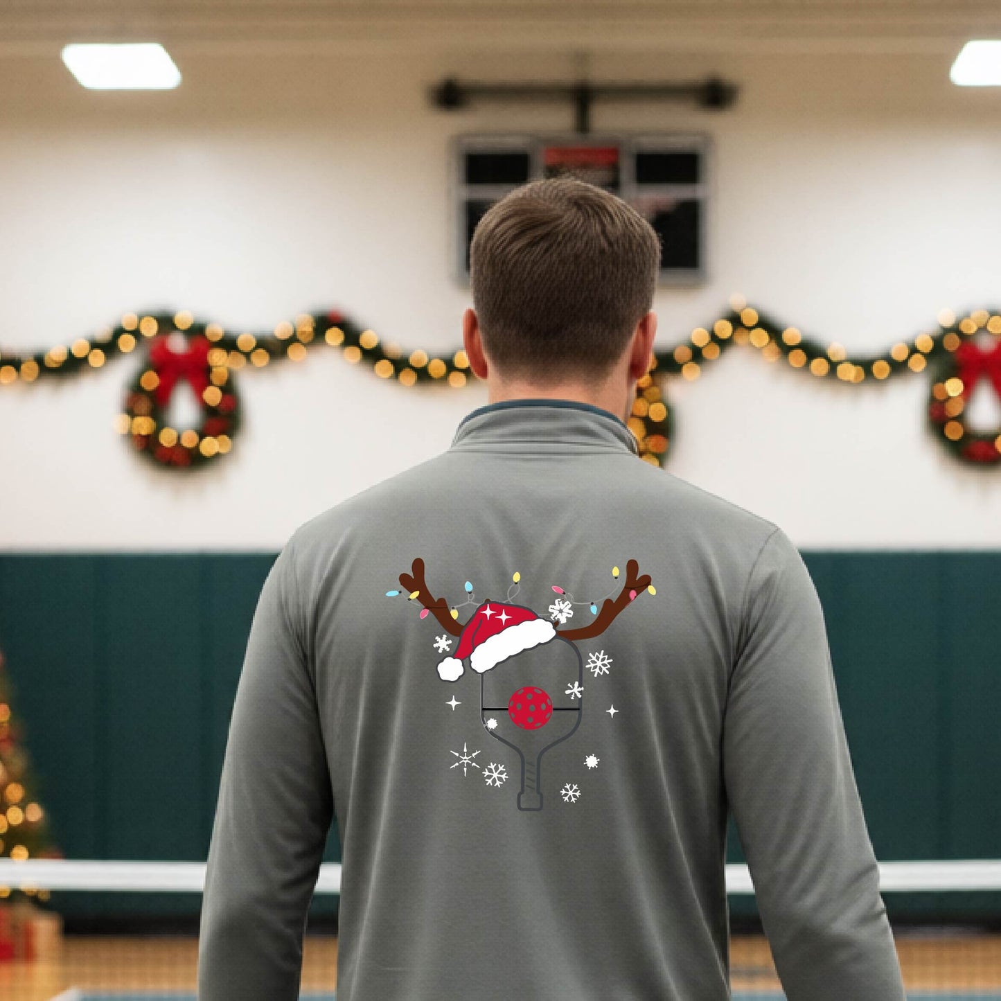 A man viewed from behind wearing a gray long-sleeve quarter-zip athletic shirt with a Christmas-themed back graphic of a pickleball paddle under a reindeer antler Santa hat. He is standing onpickleball court with holiday decorations.