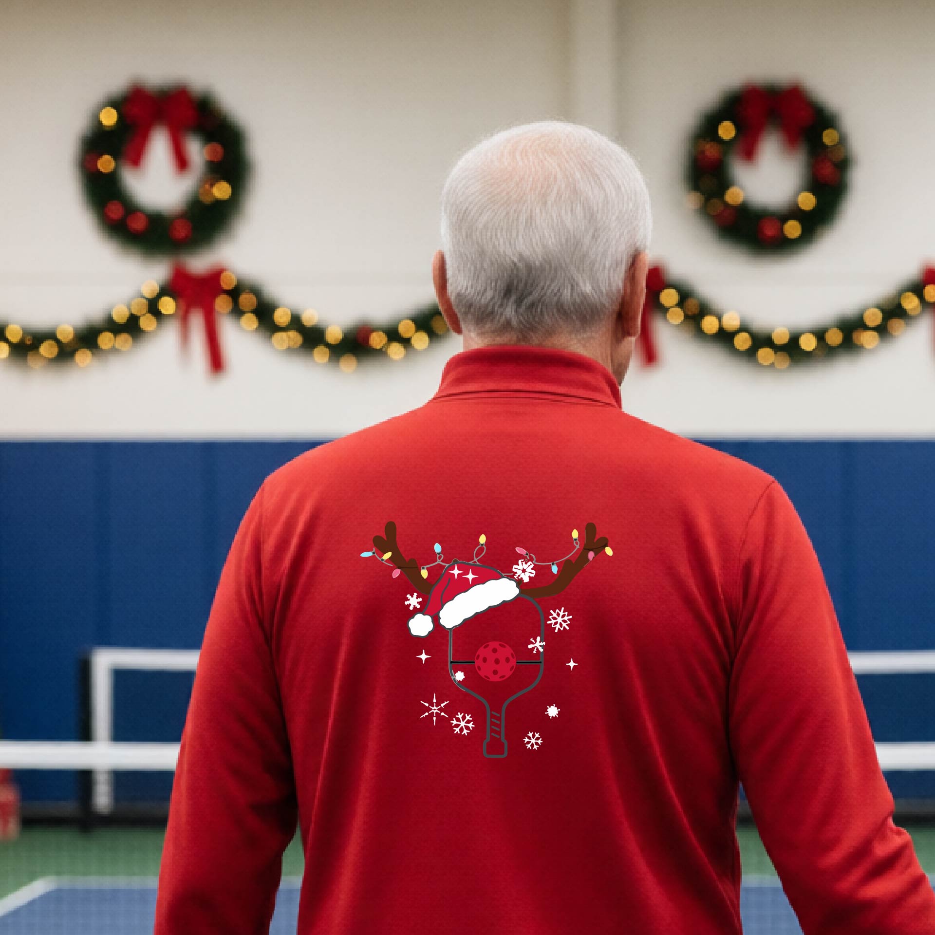 A man viewed from behind wearing a red long-sleeve quarter-zip athletic shirt with a Christmas-themed back graphic of a pickleball paddle under a reindeer antler Santa hat. He is standing onpickleball court with holiday decorations.